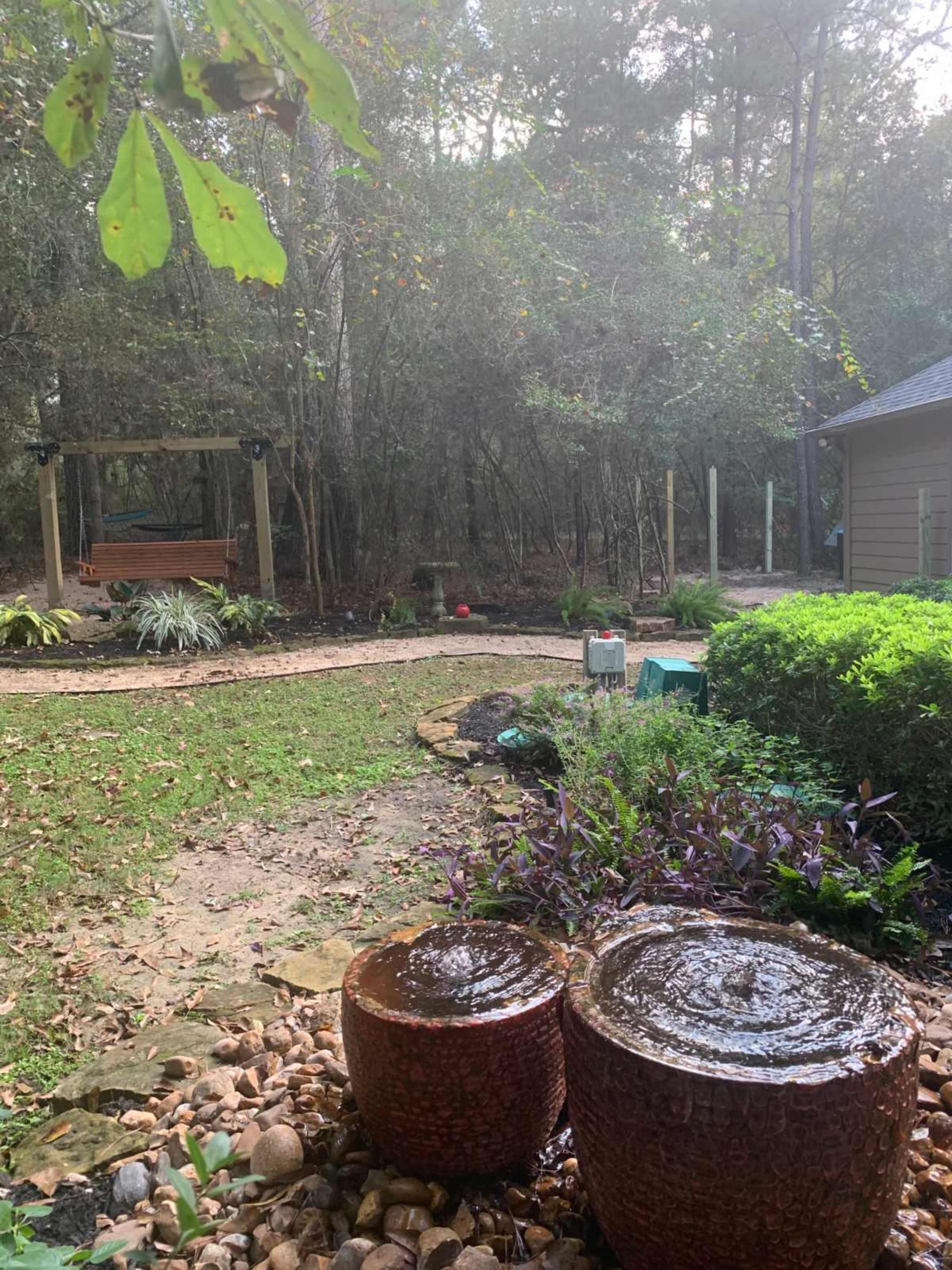 A landscaped backyard with two water fountains in the foreground, a swing in the background, and surrounding lush greenery.