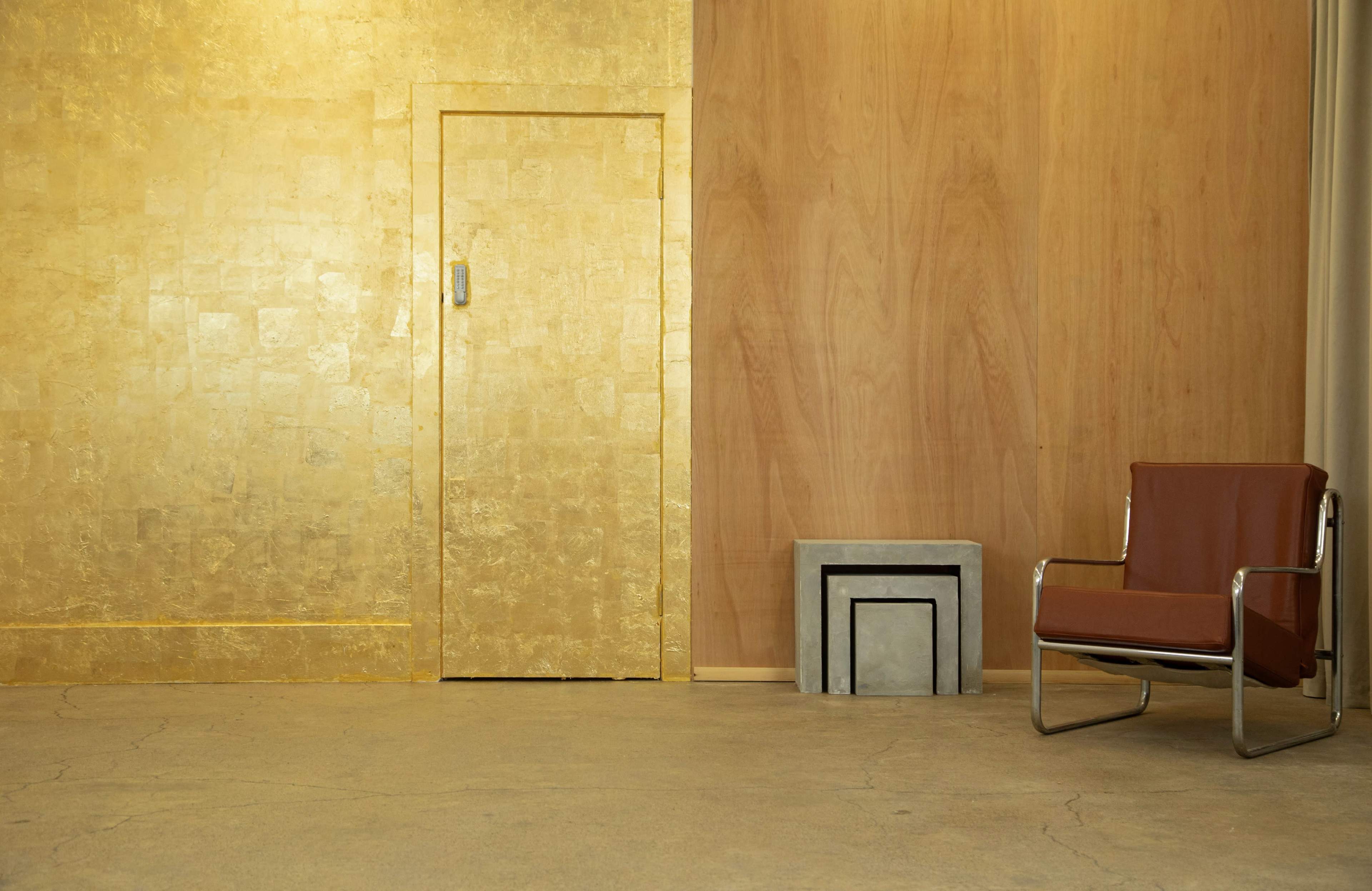 The scene features a minimalist interior with a gold wall, a wooden wall, a brown chair, and a concrete cube.
