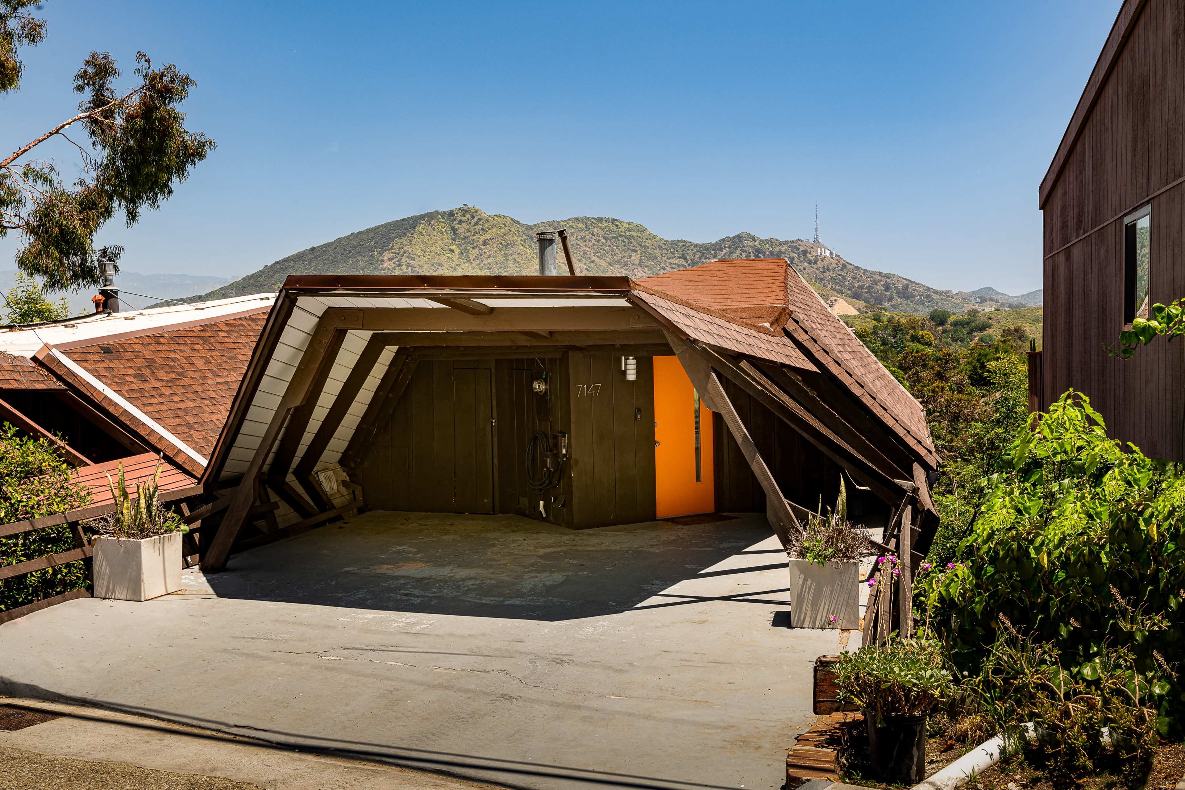 A unique A-frame house with a sloped roof and an orange door, set against a backdrop of mountains and a clear blue sky.