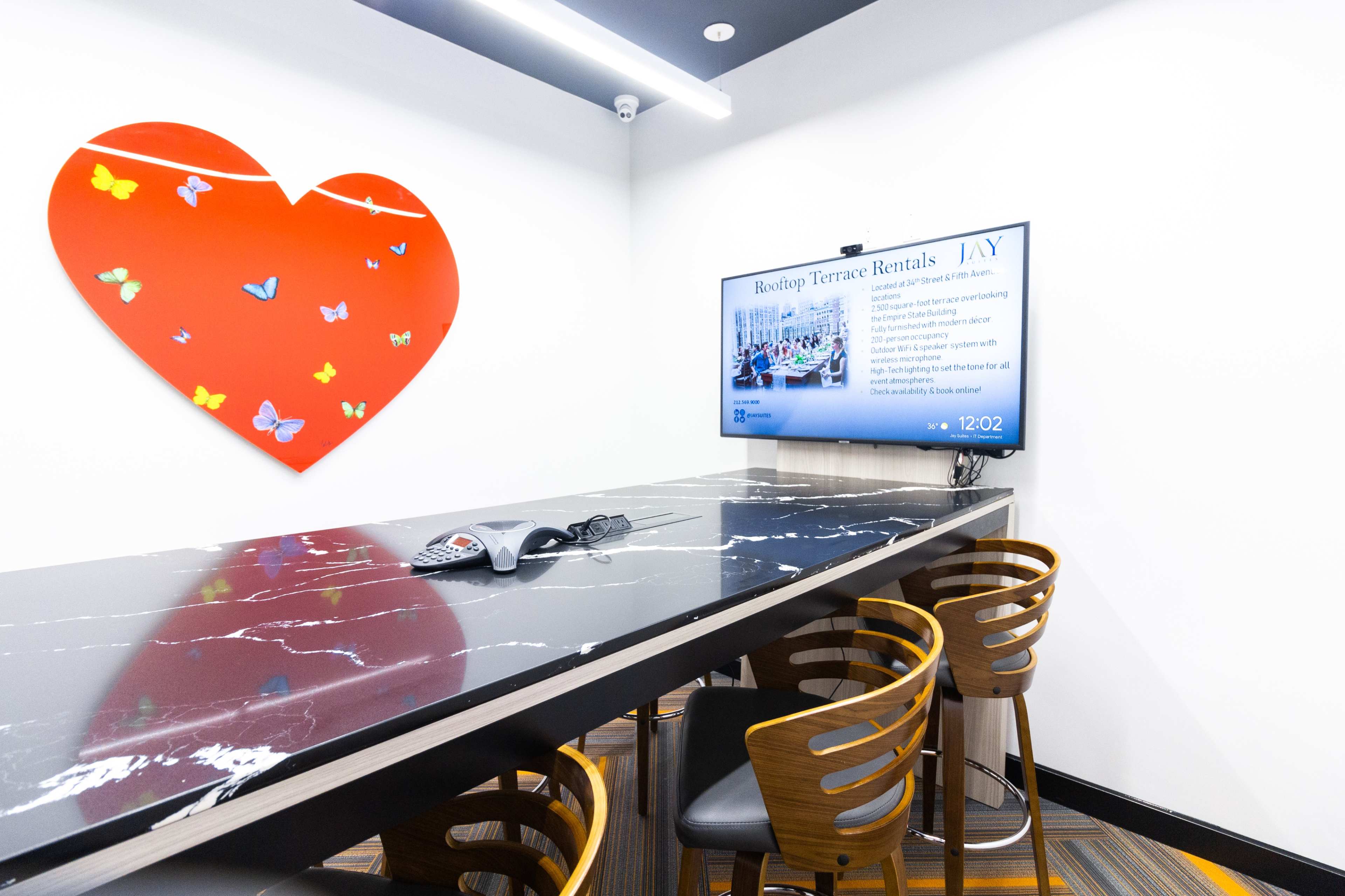A conference room features a large black marble table, wooden chairs, a television displaying information about terrace rentals, and a heart-shaped artwork on the wall.