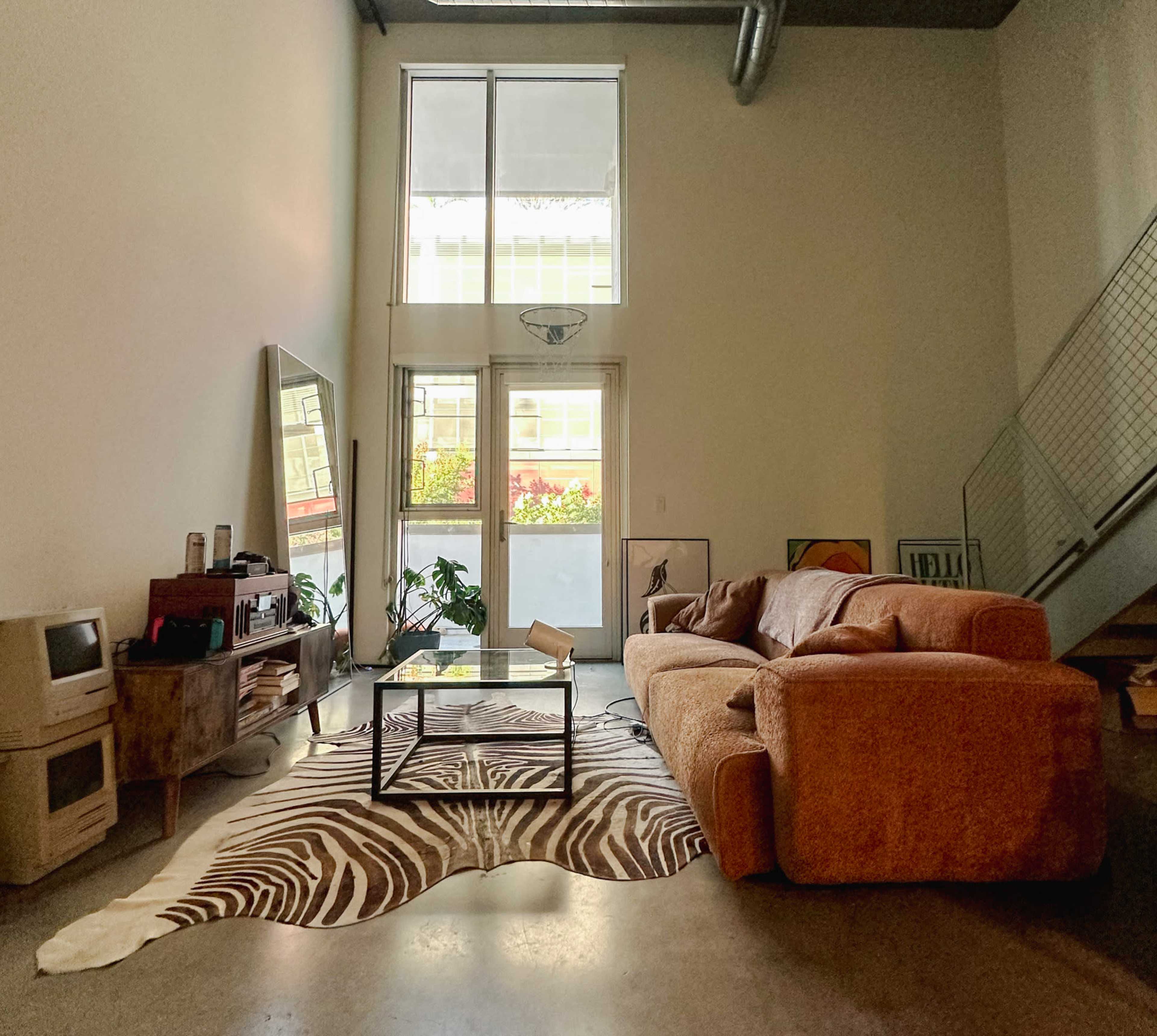 A spacious living area features a large orange couch, a glass coffee table on a zebra-print rug, and a mirror against a wall with large windows allowing natural light.