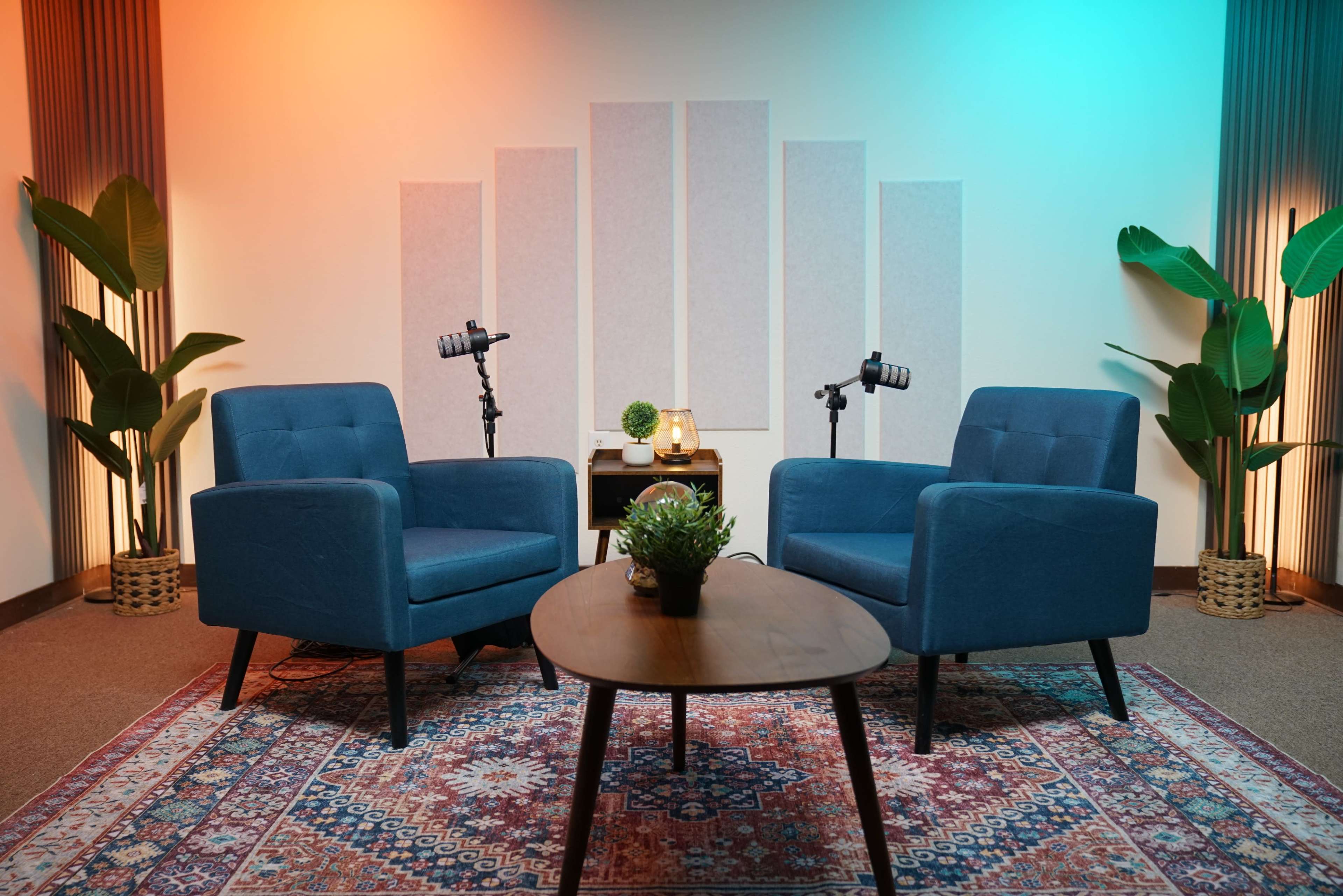 A cozy interview setup featuring two blue chairs, a small table with decorative items, and plants, all under soft, colored lighting.