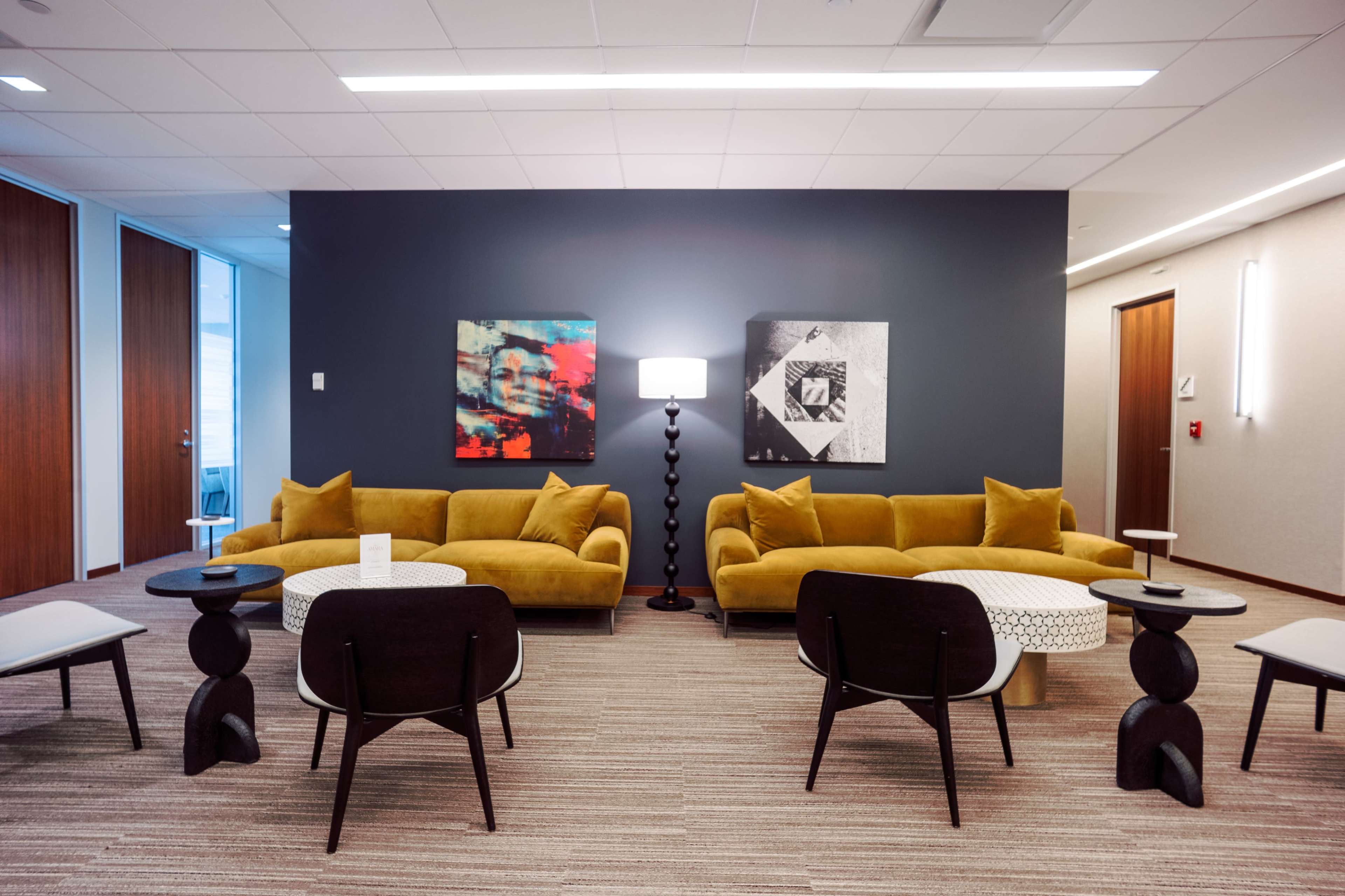 The image depicts a modern lounge area featuring two mustard-colored sofas, two small circular tables, and two artworks on the wall, along with a standing lamp.