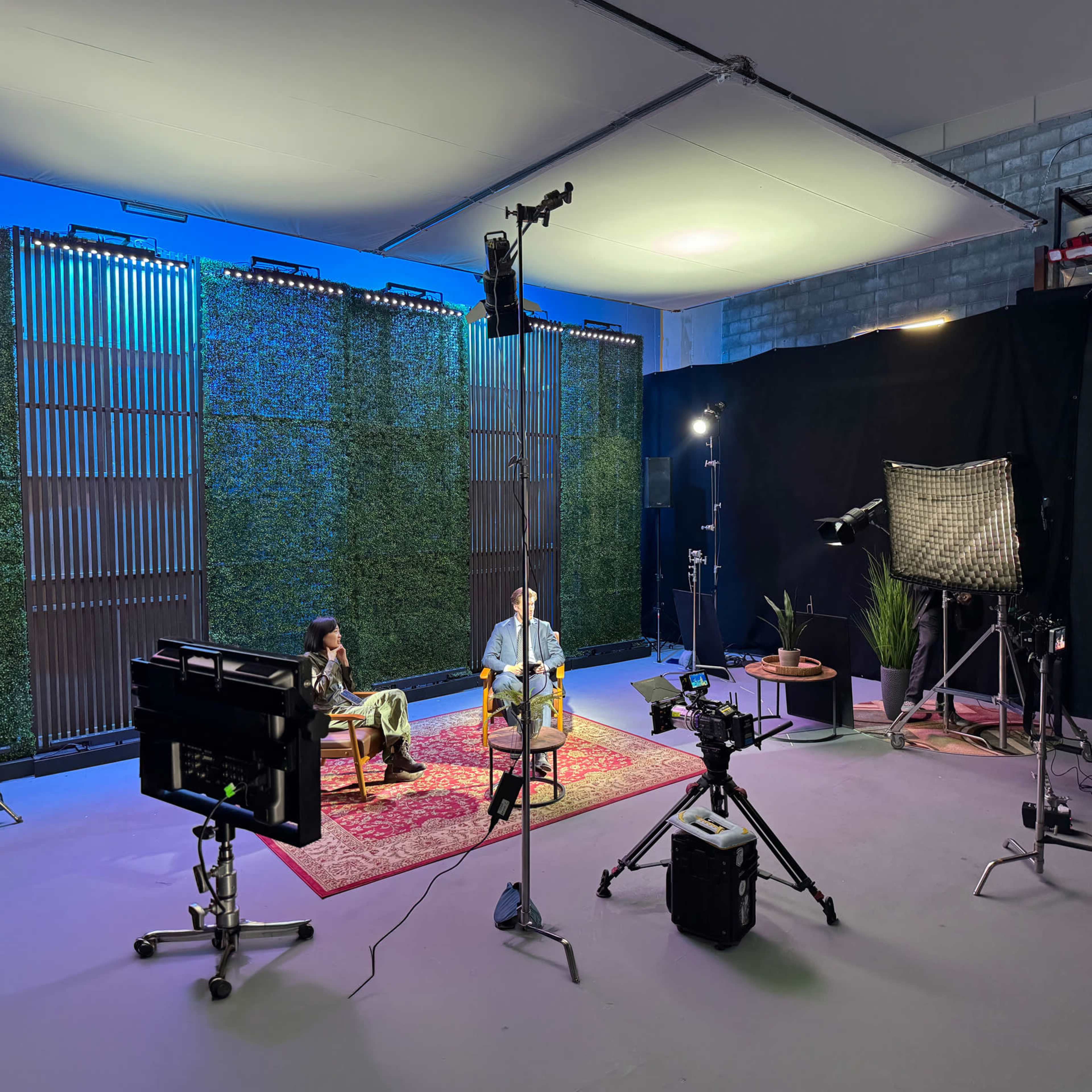 The image shows a filming studio setup with two people seated on chairs in front of a backdrop of greenery, illuminated by various studio lights and cameras positioned around them.