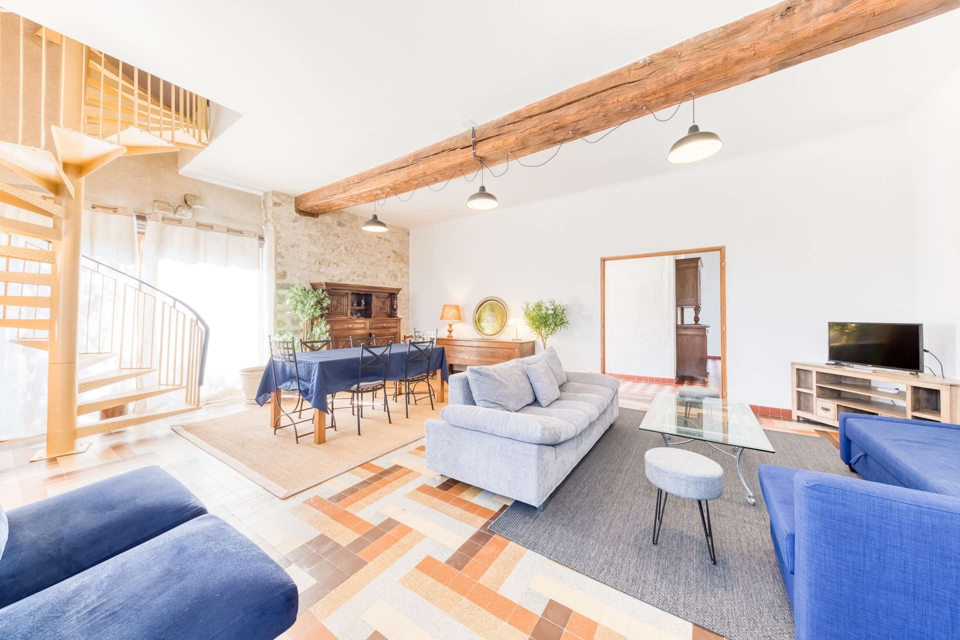 The image shows a spacious living room with a blue sofa, a glass coffee table, a dining area with a dark table and chairs, and a spiral staircase in the background.