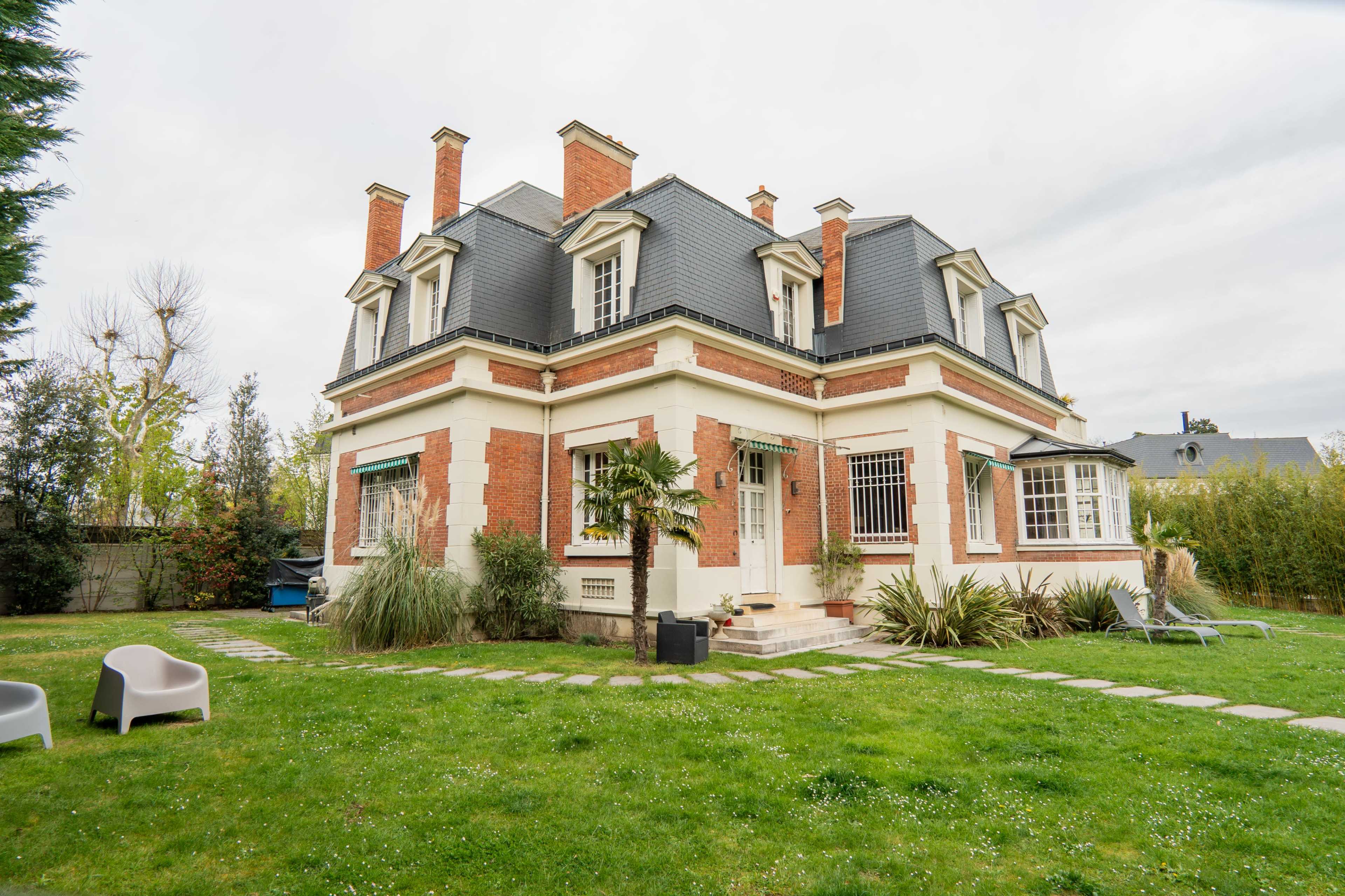 A large house with a gray roof and red brick walls is surrounded by a green lawn and landscaping.