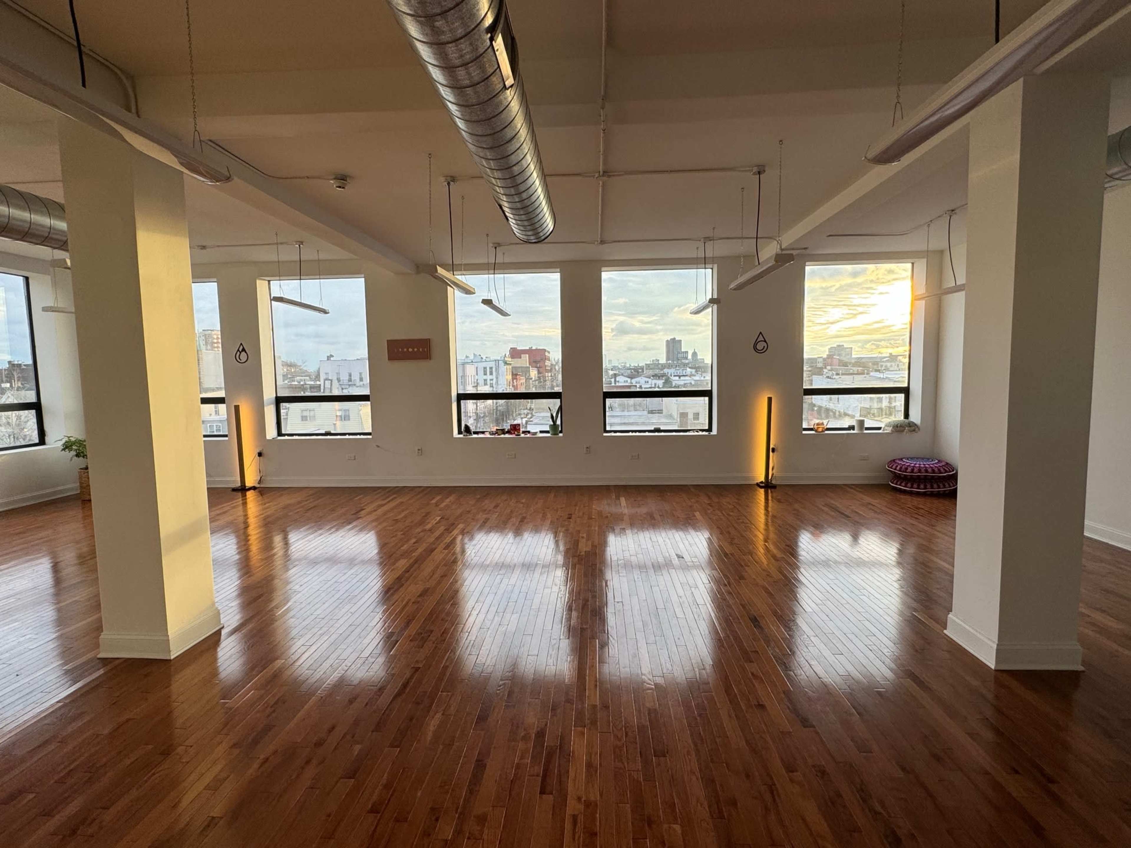 The image shows a spacious, empty room with polished wooden floors and large windows offering a view of the city skyline at sunset.