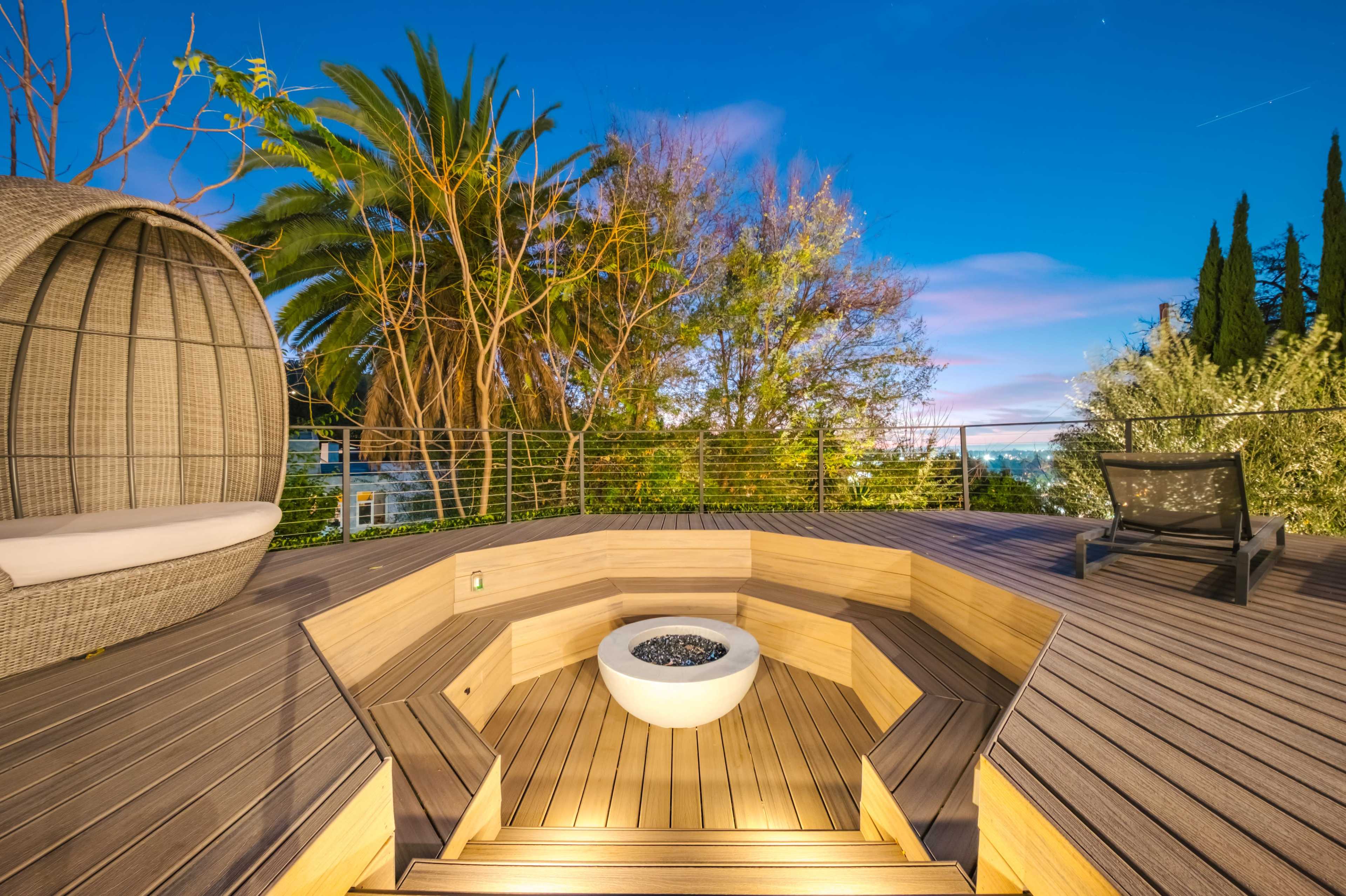 The image shows a modern outdoor deck featuring a circular fire pit surrounded by wooden seating areas, with lush greenery and palm trees in the background.