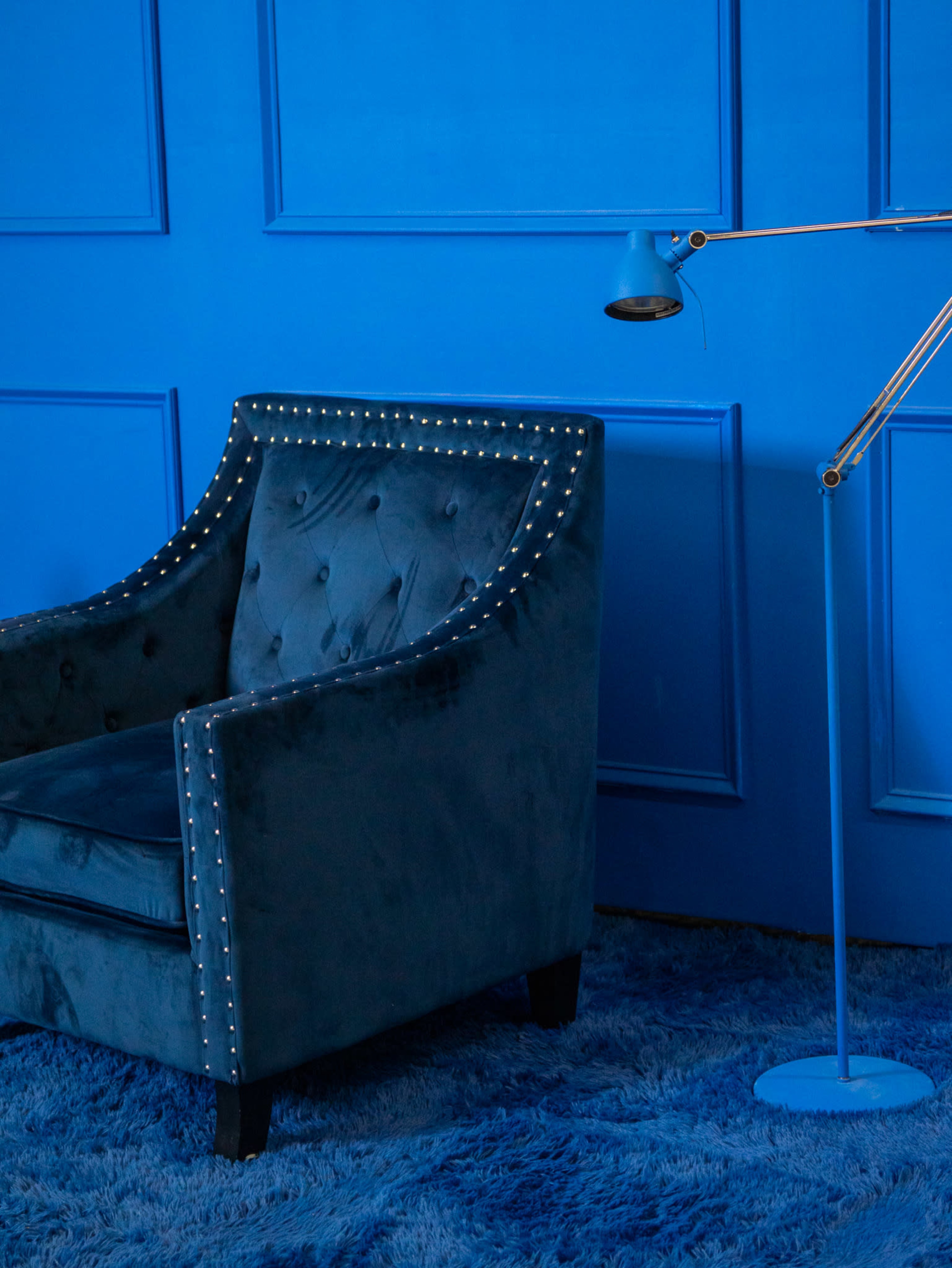 A tufted blue armchair with metallic studs sits beside a blue floor lamp against a blue paneled wall.