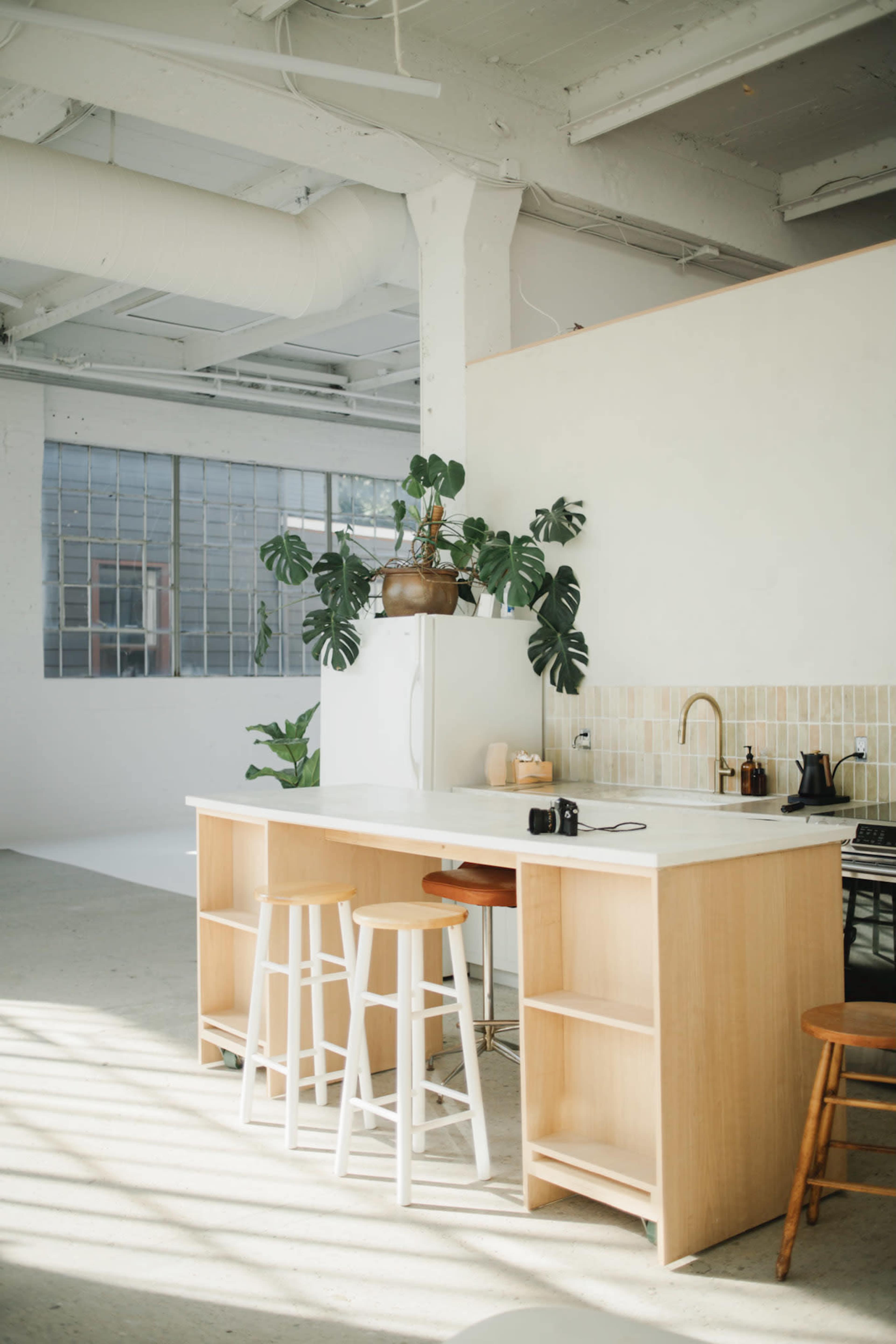 A modern kitchen with a light wood island, two stools, plants, and a minimalist design.