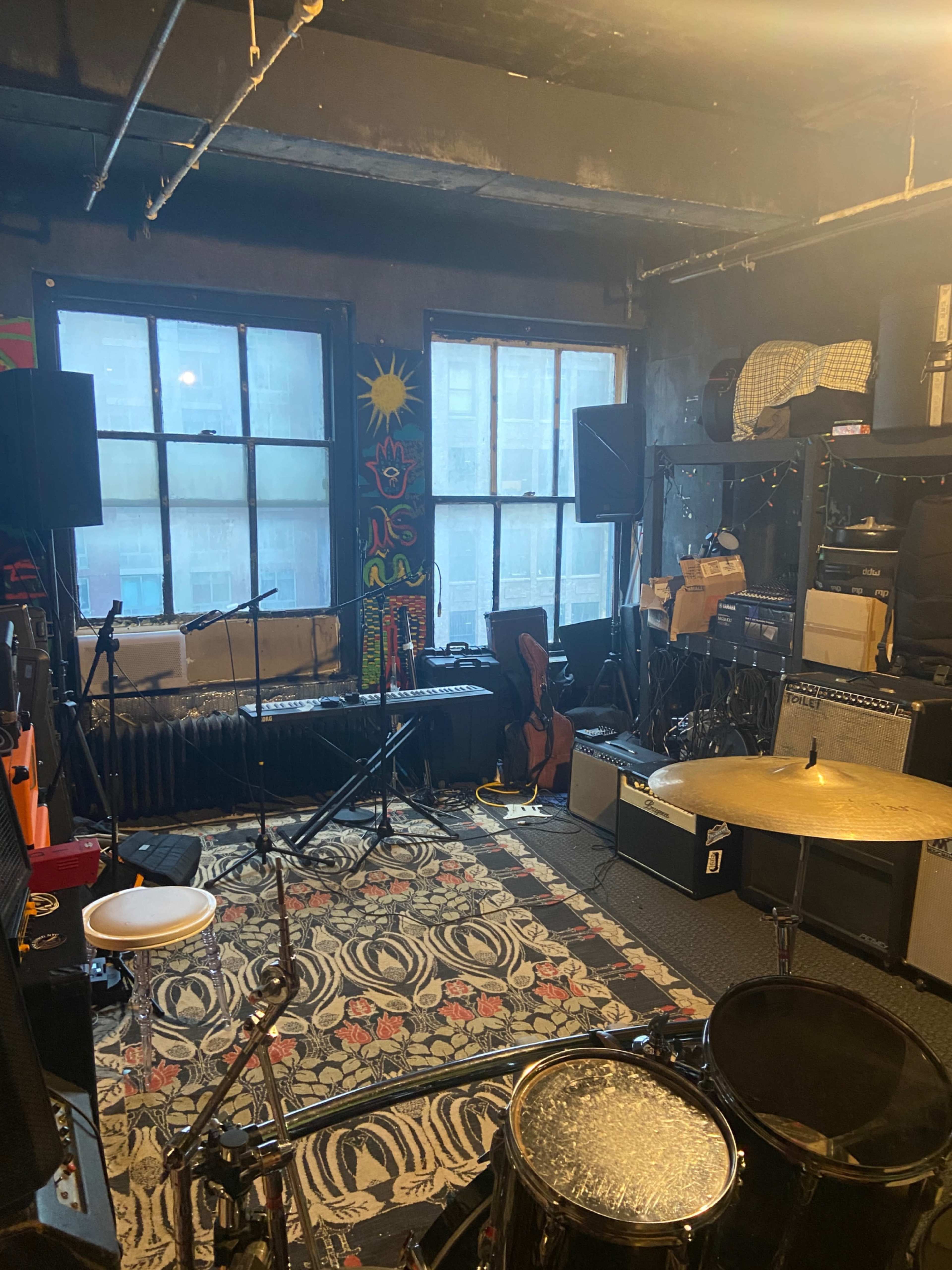 The image shows a music practice space with instruments including a drum set, amplifiers, and a keyboard, surrounded by windows and colorful wall decorations.