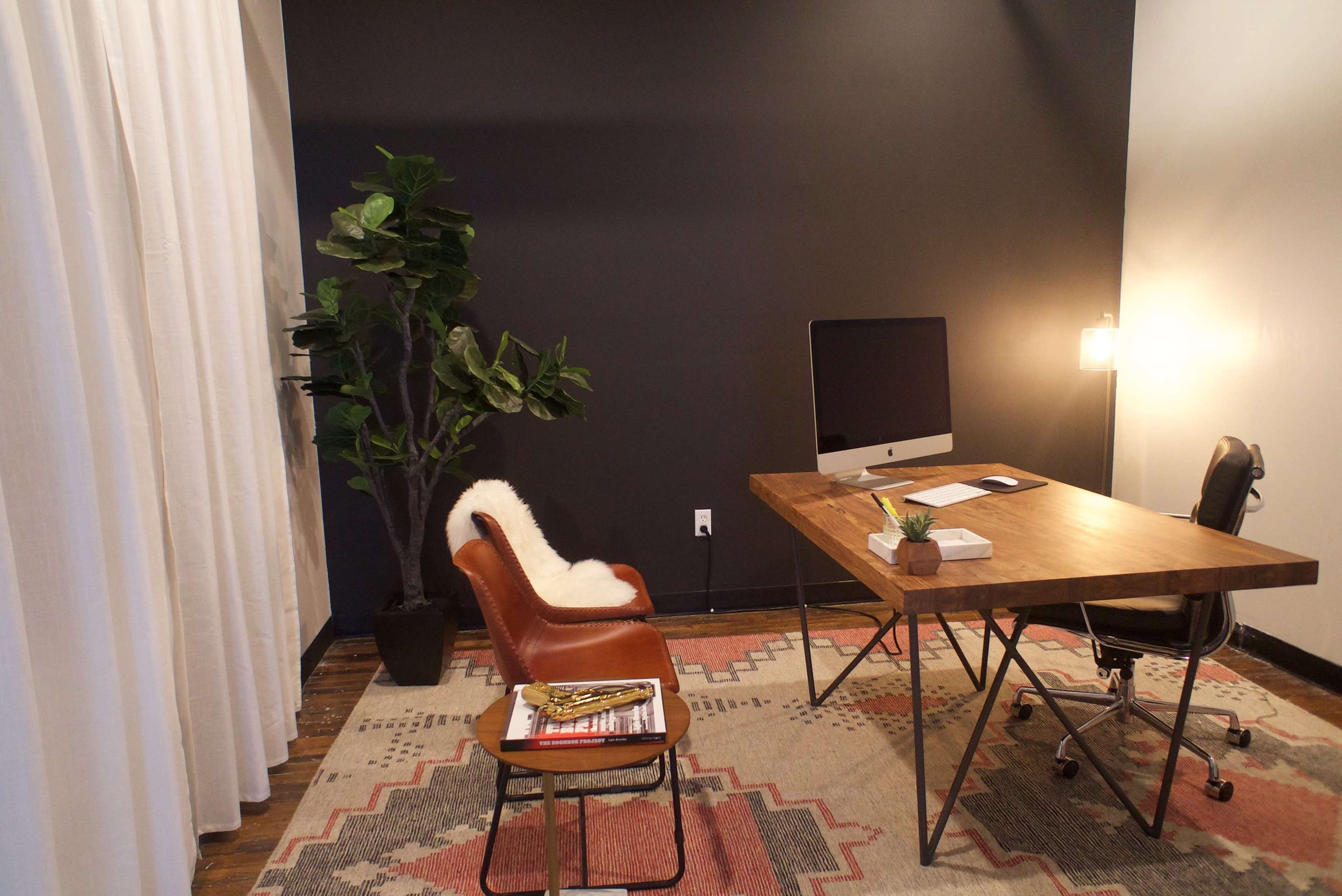 The image shows a minimalist office space with a wooden desk, an iMac, a brown leather chair, a potted plant, and a patterned rug on the floor.