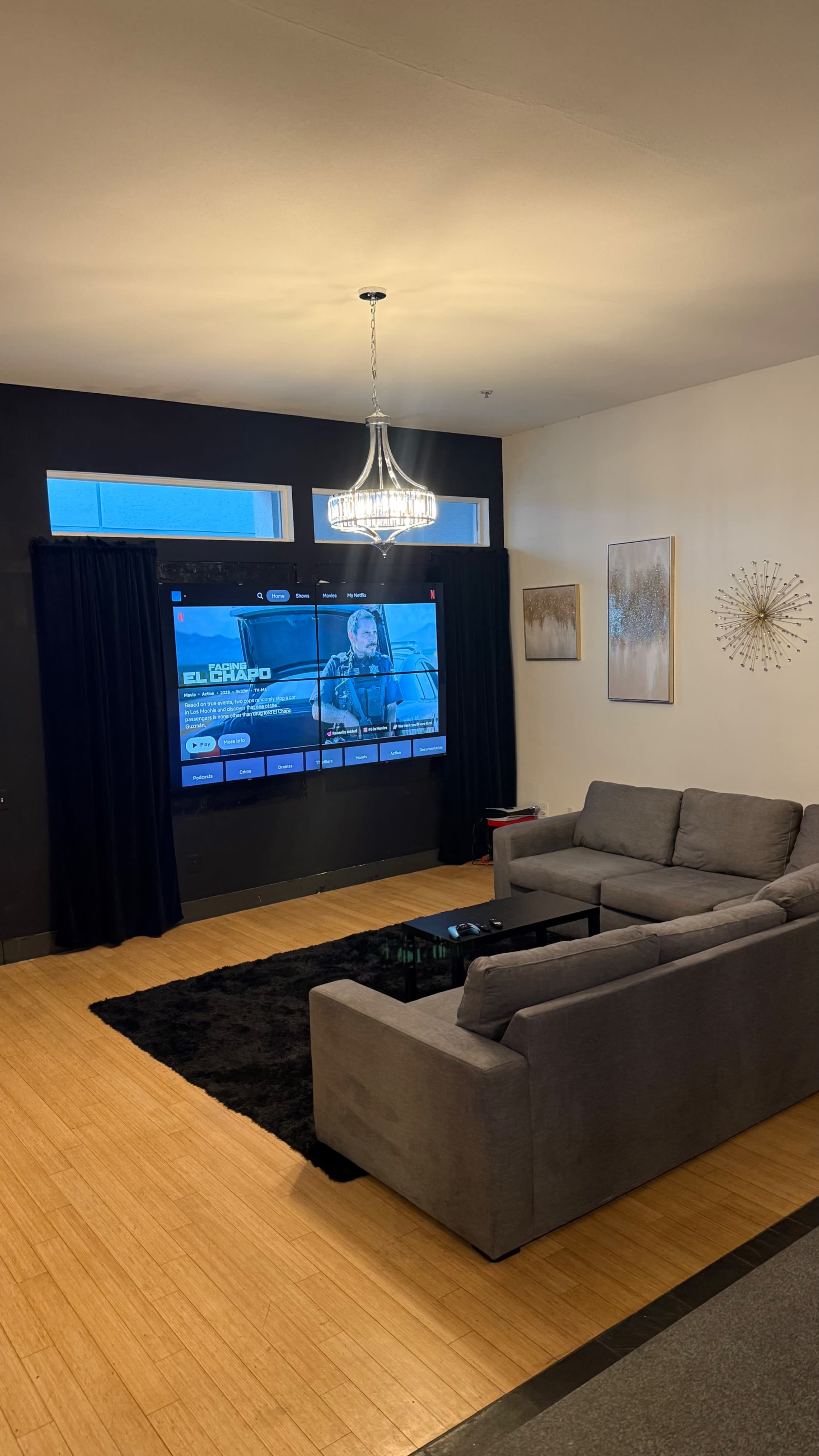 A cozy living room features a gray couch and coffee table facing a large TV screen displaying a streaming interface, with decorative artwork on the walls and a chandelier overhead.