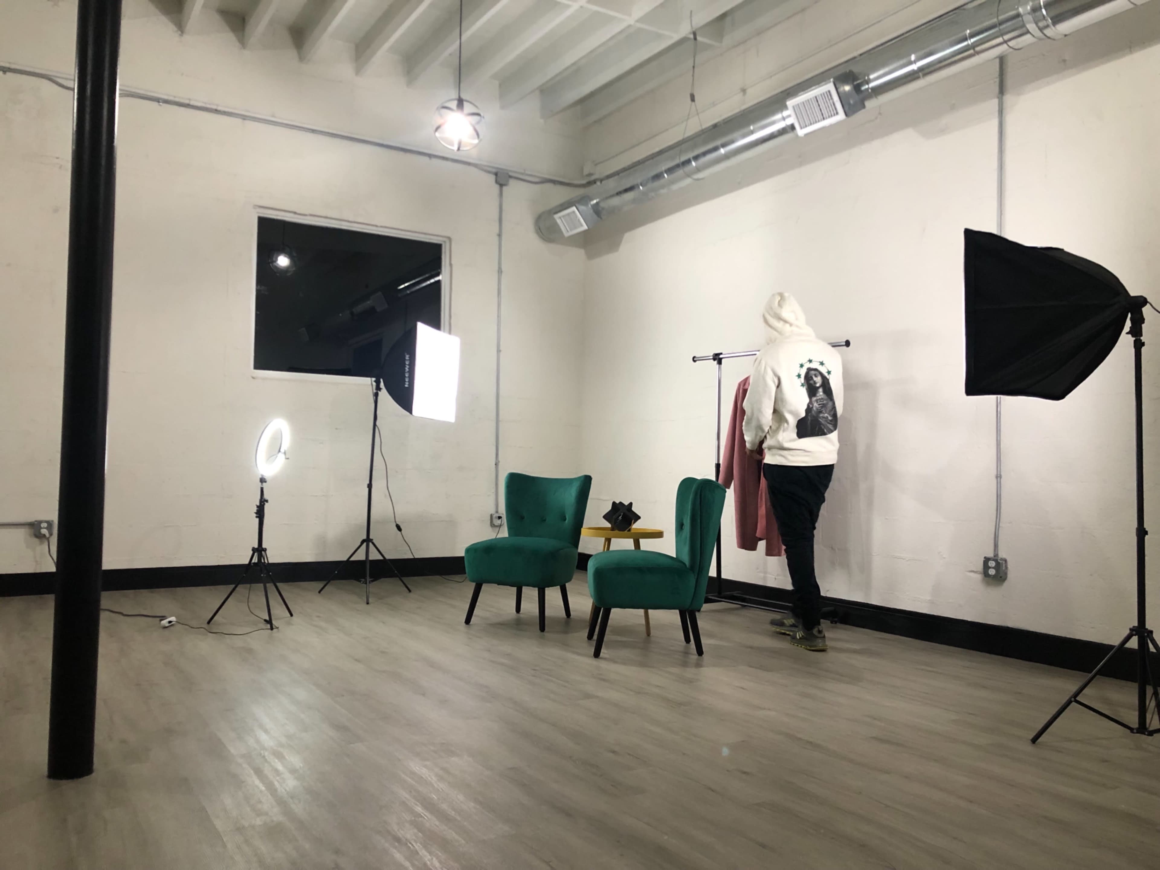 A person in a hoodie arranges a clothing item on a rack in a well-lit studio with green chairs and studio lighting equipment.