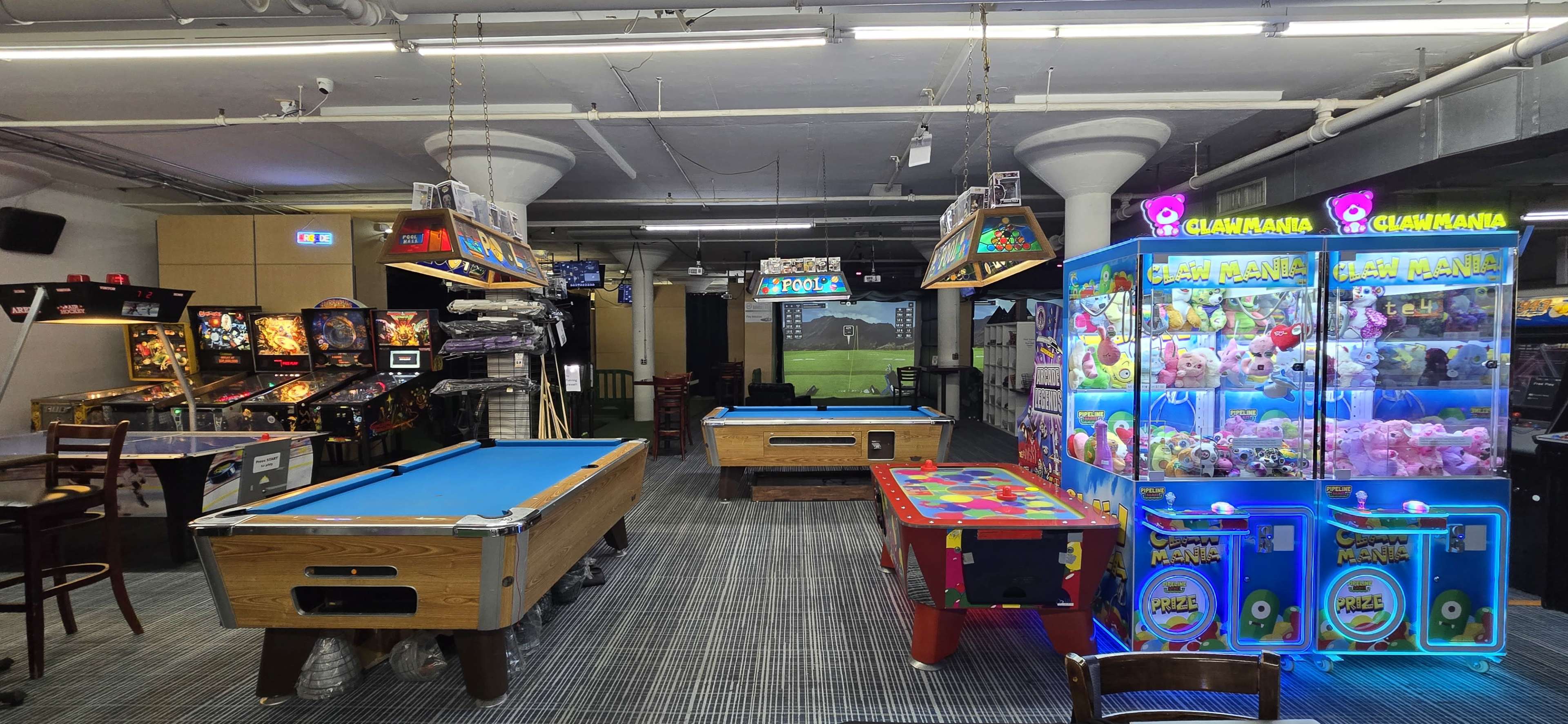 The image shows an arcade with various games, including pinball machines, a pool table, and a claw machine, set in a spacious room with overhead lighting.