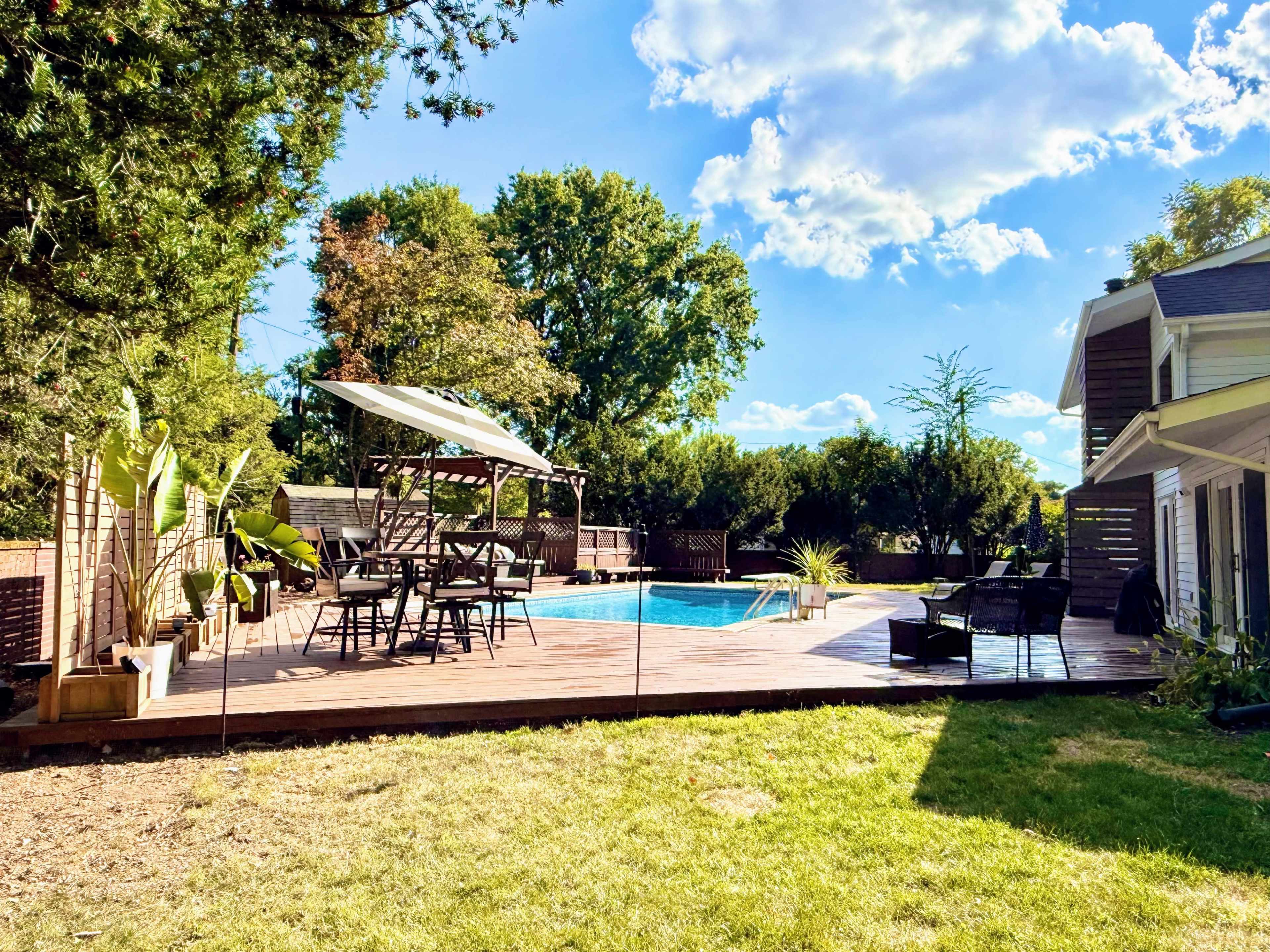 The image shows a backyard with a wooden deck, a swimming pool, and seating areas surrounded by greenery and trees under a blue sky with white clouds.