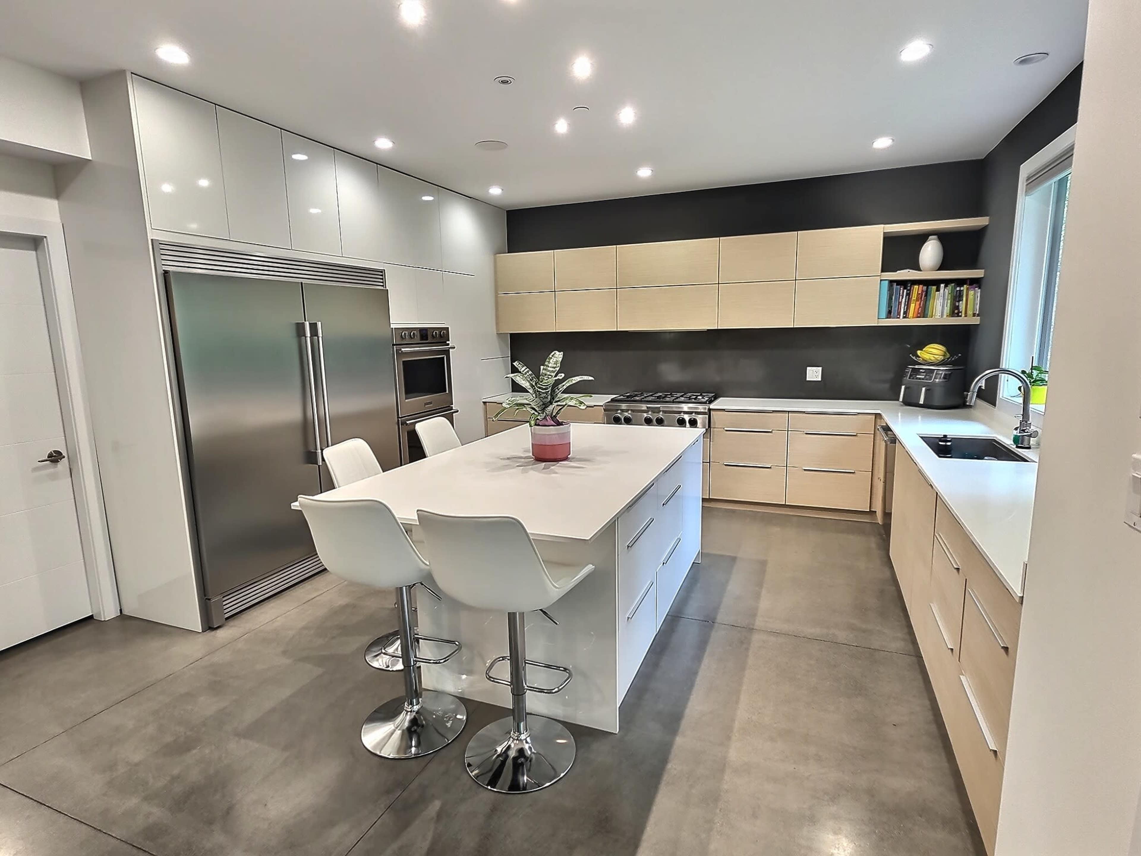 A modern kitchen features a central island with bar stools, sleek cabinetry, stainless steel appliances, and a large window.