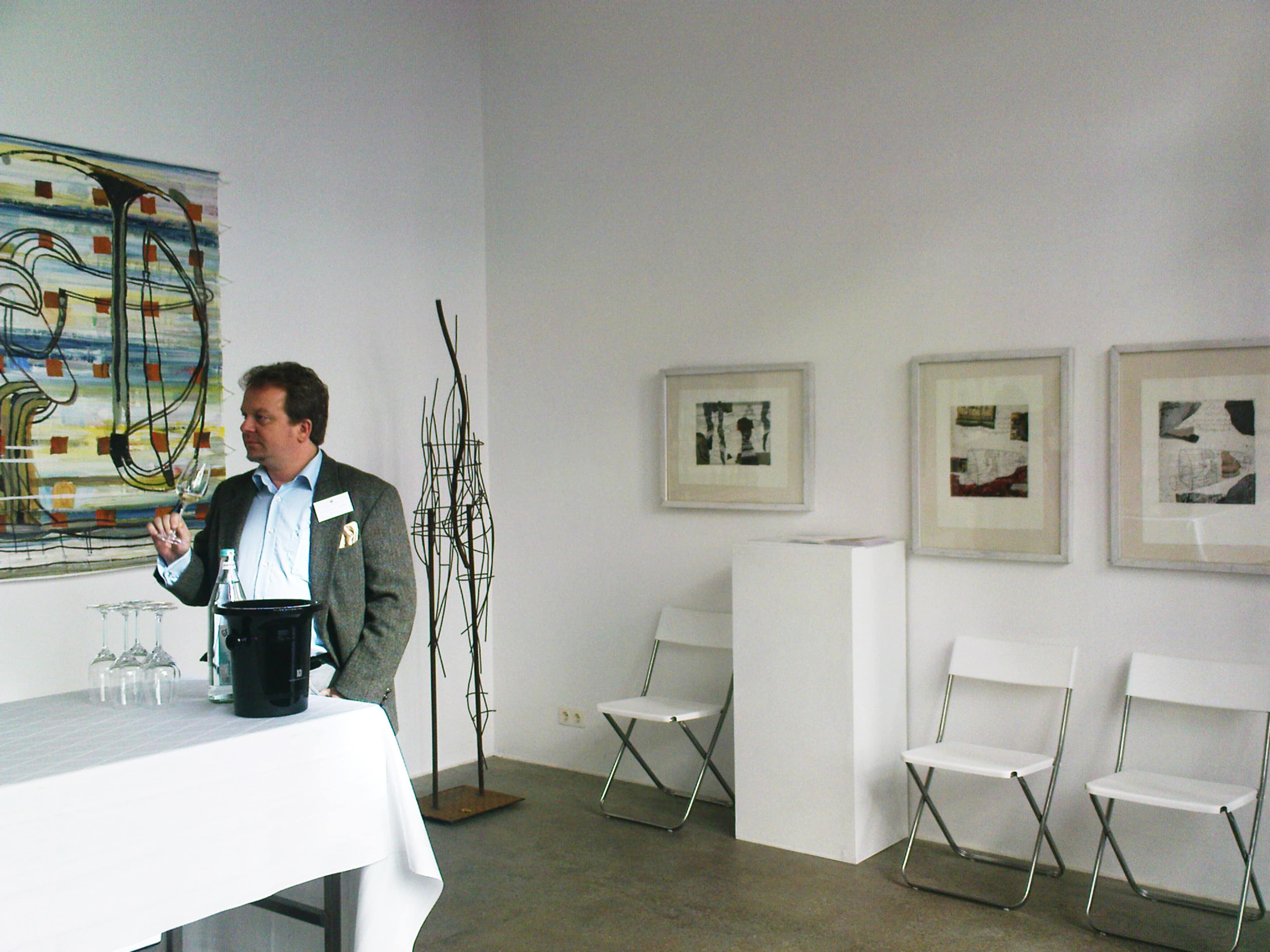 A man stands behind a white table displaying glasses and a black container, with abstract art on the wall and empty folding chairs nearby.