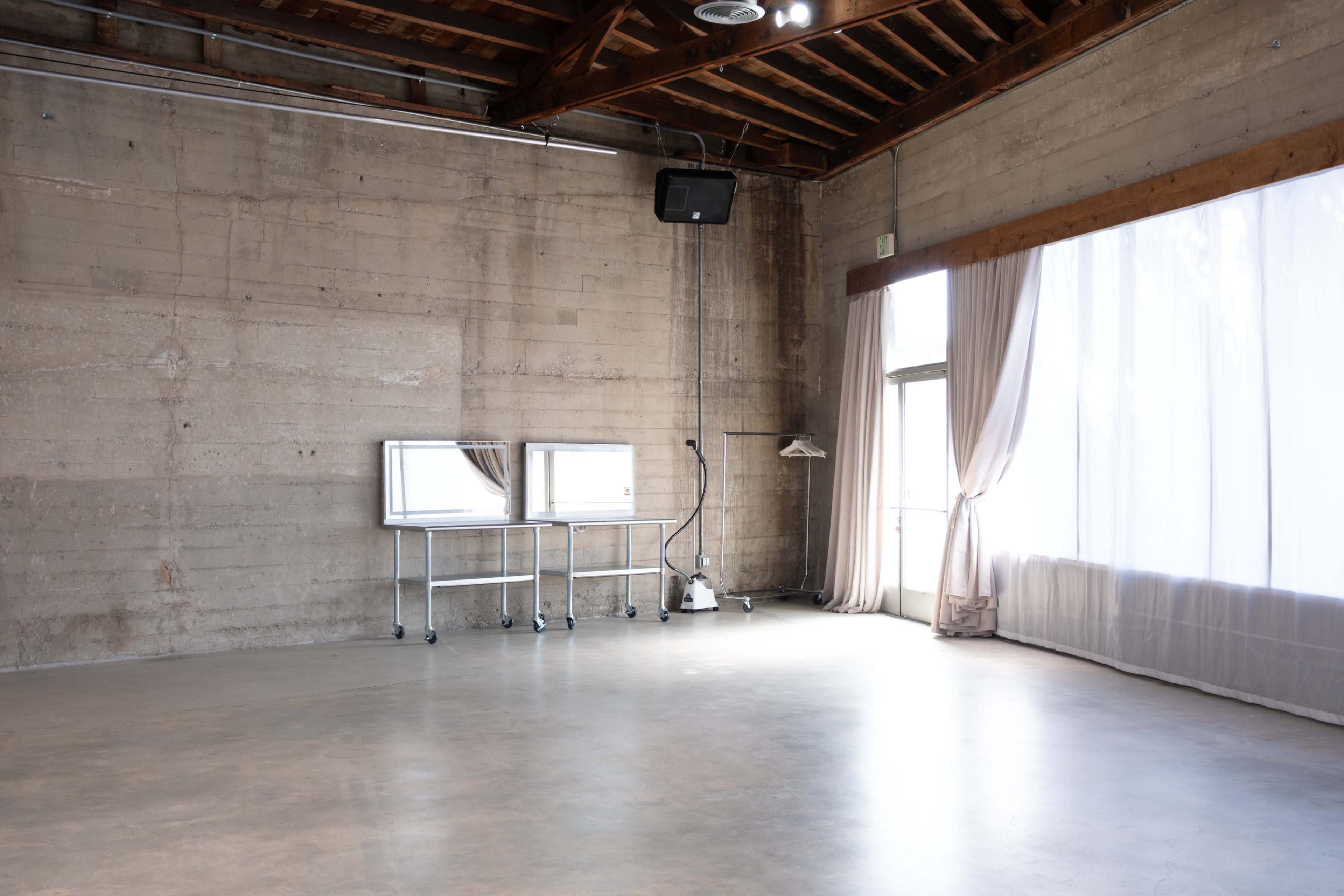 The image shows a spacious, minimalist room with concrete walls, large windows covered by sheer curtains, and two mirrors on a metal stand.
