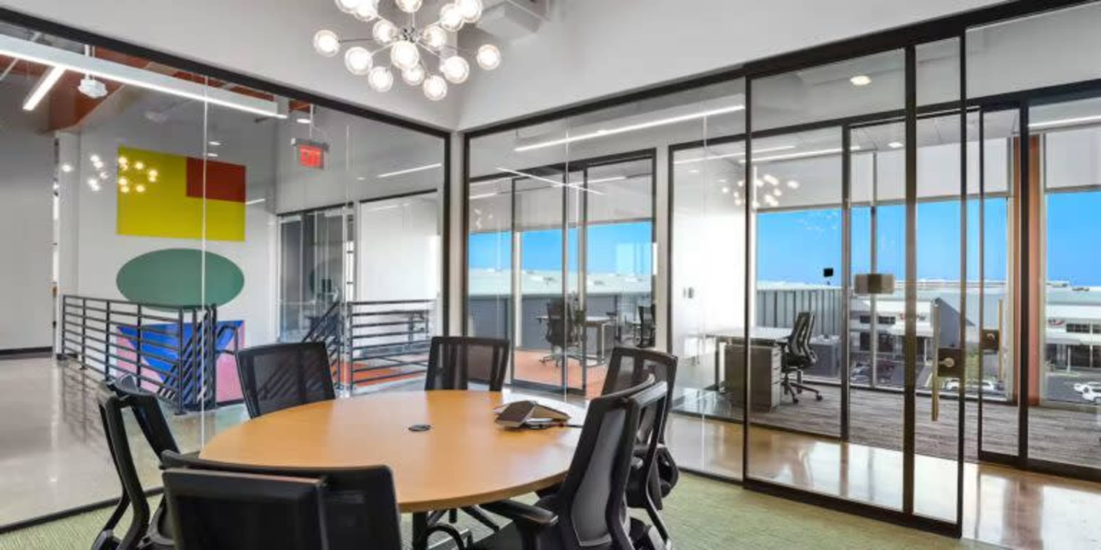 The image shows a modern office space with a round conference table surrounded by black chairs, and large glass panels separating different work areas.
