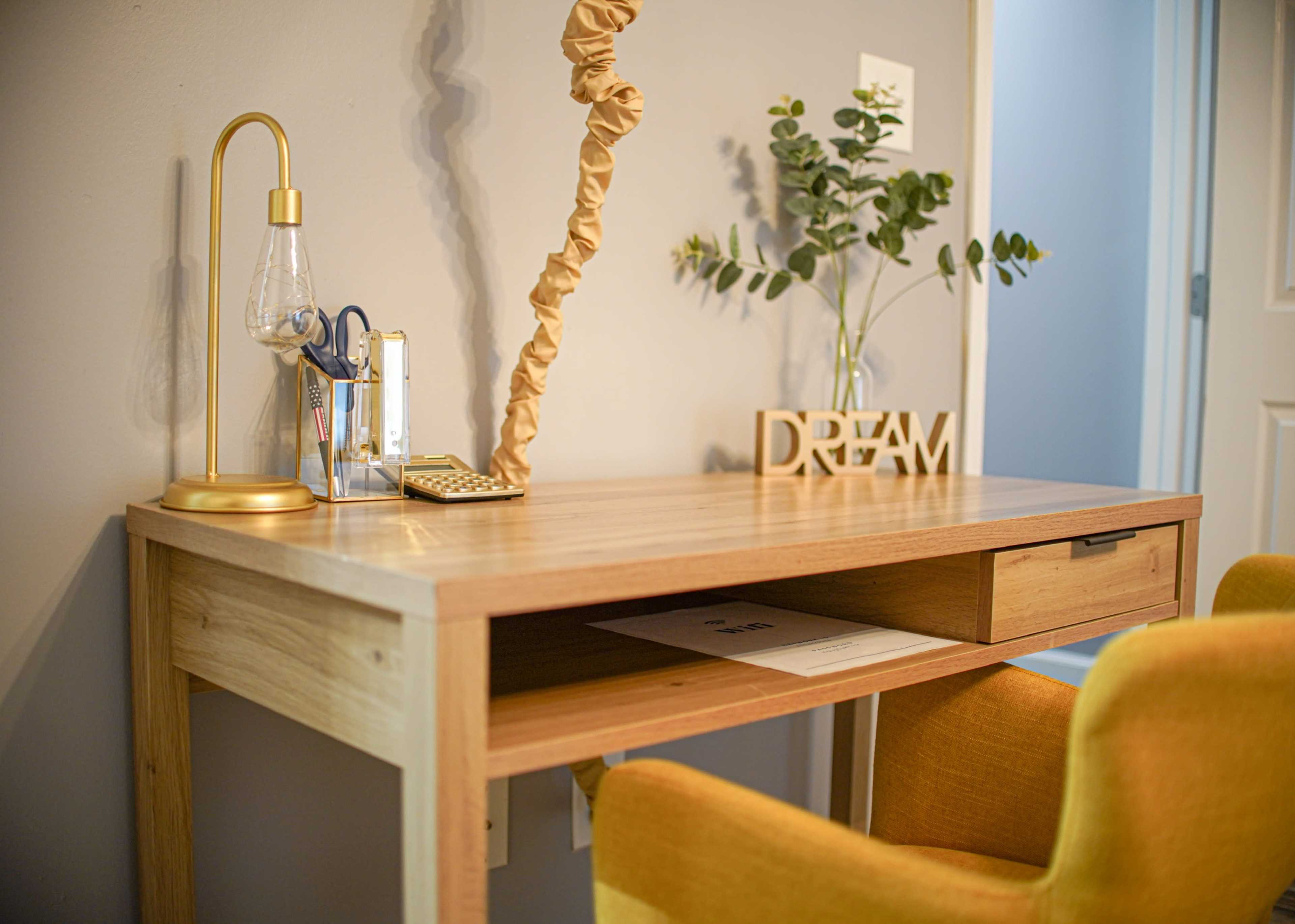 A wooden desk is styled with a lamp, a small plant, and a decorative "DREAM" sign beside a yellow chair.