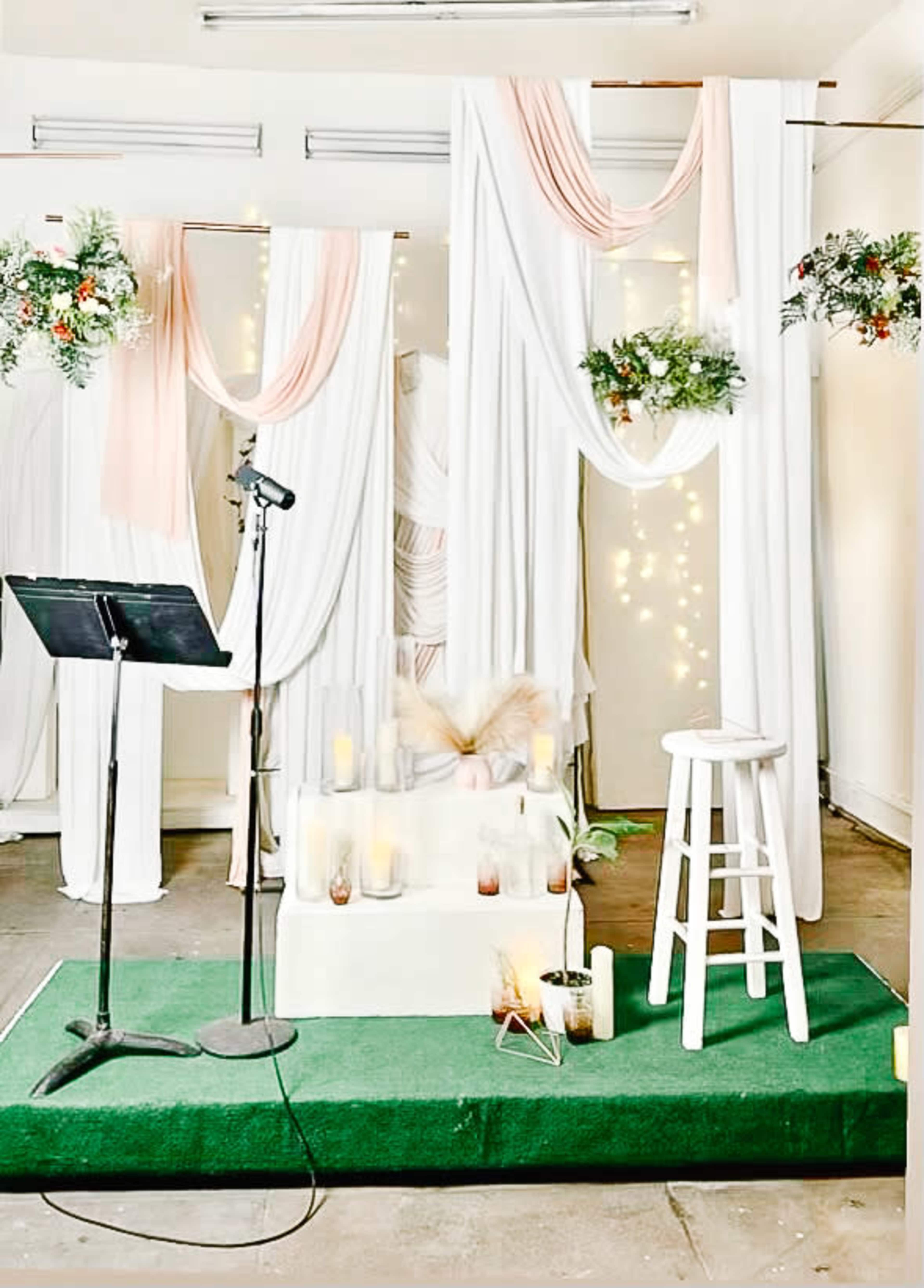 The image shows a decorated stage with white drapes, greenery, candles, a microphone stand, and a stool, set up for an event.