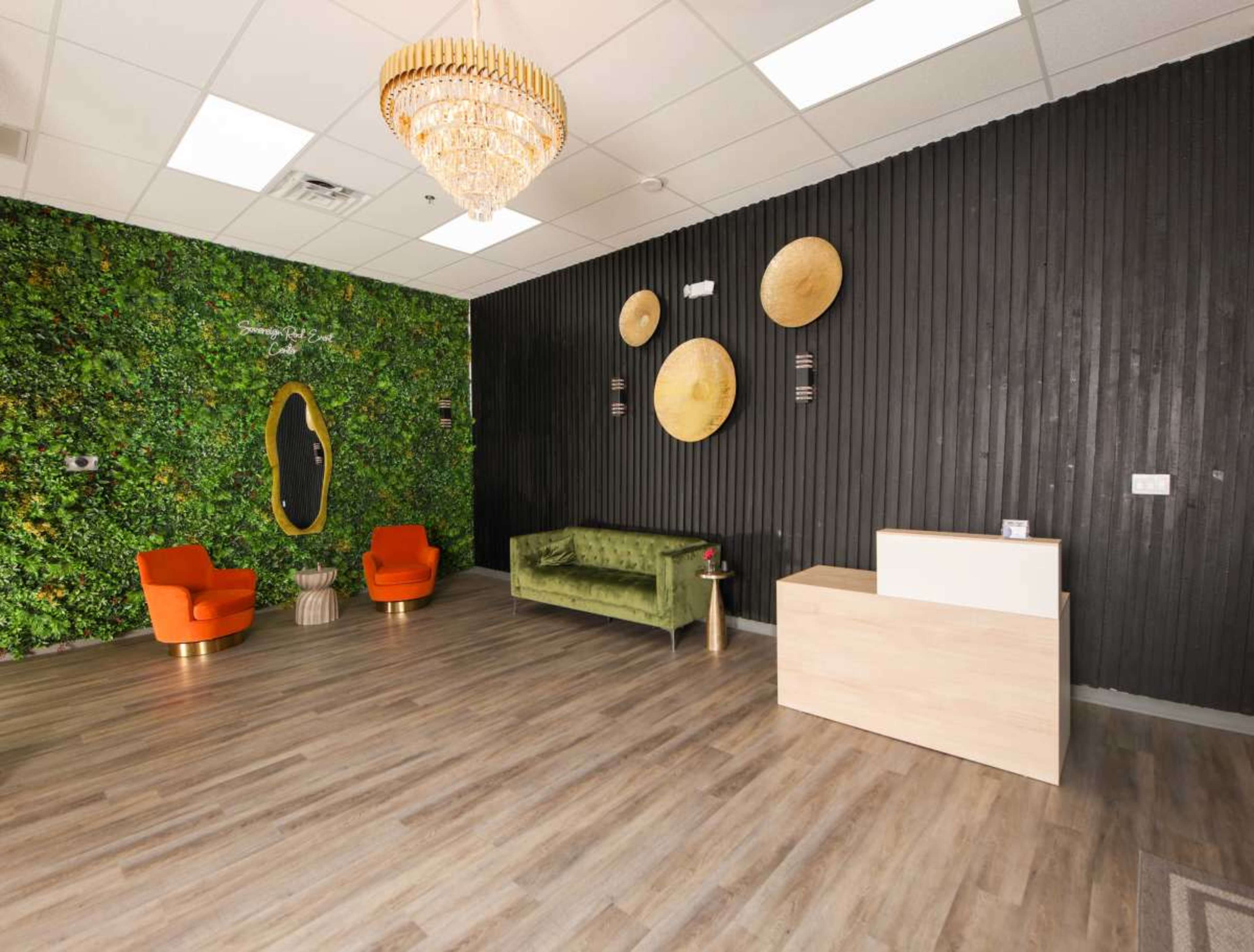 The image shows a modern interior layout featuring a green wall with plants, two orange chairs, a green sofa, and a reception desk.