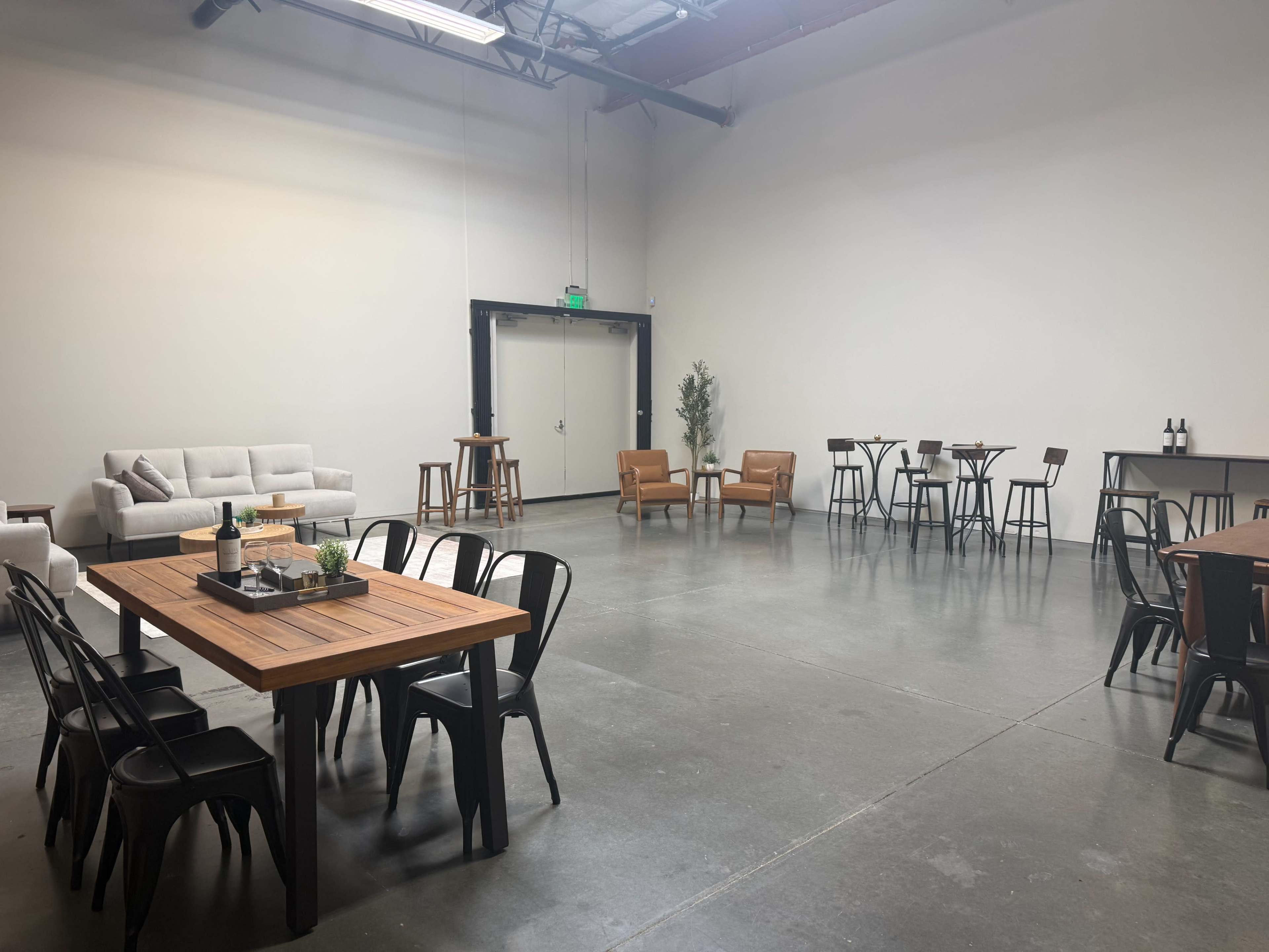 The image shows a spacious, empty industrial-style room with wooden furniture, including tables and chairs, and a seating area with a sofa and armchairs.