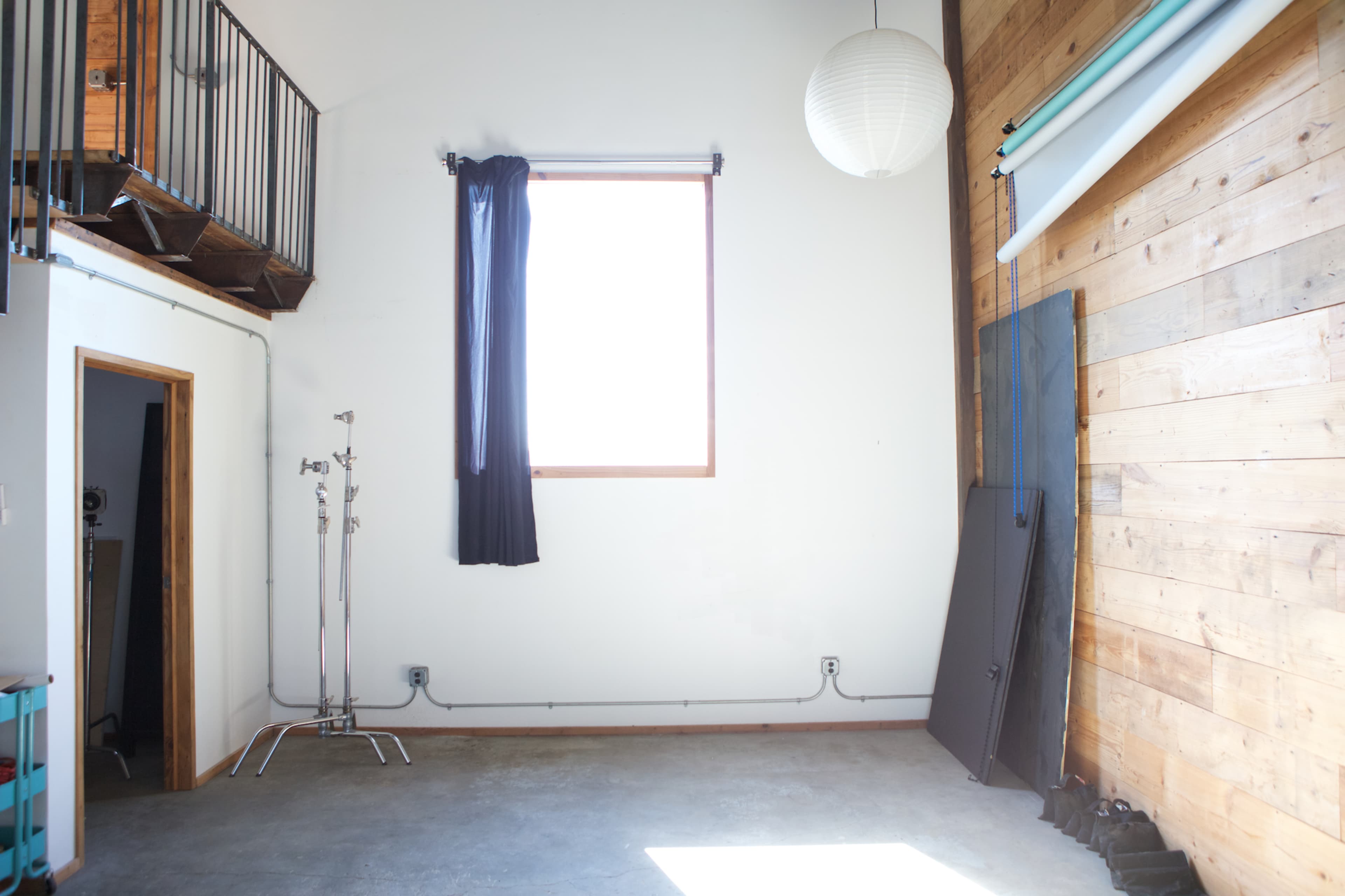 The image shows a minimalist interior space with a large window, wooden wall panels, and a few studio equipment stands.