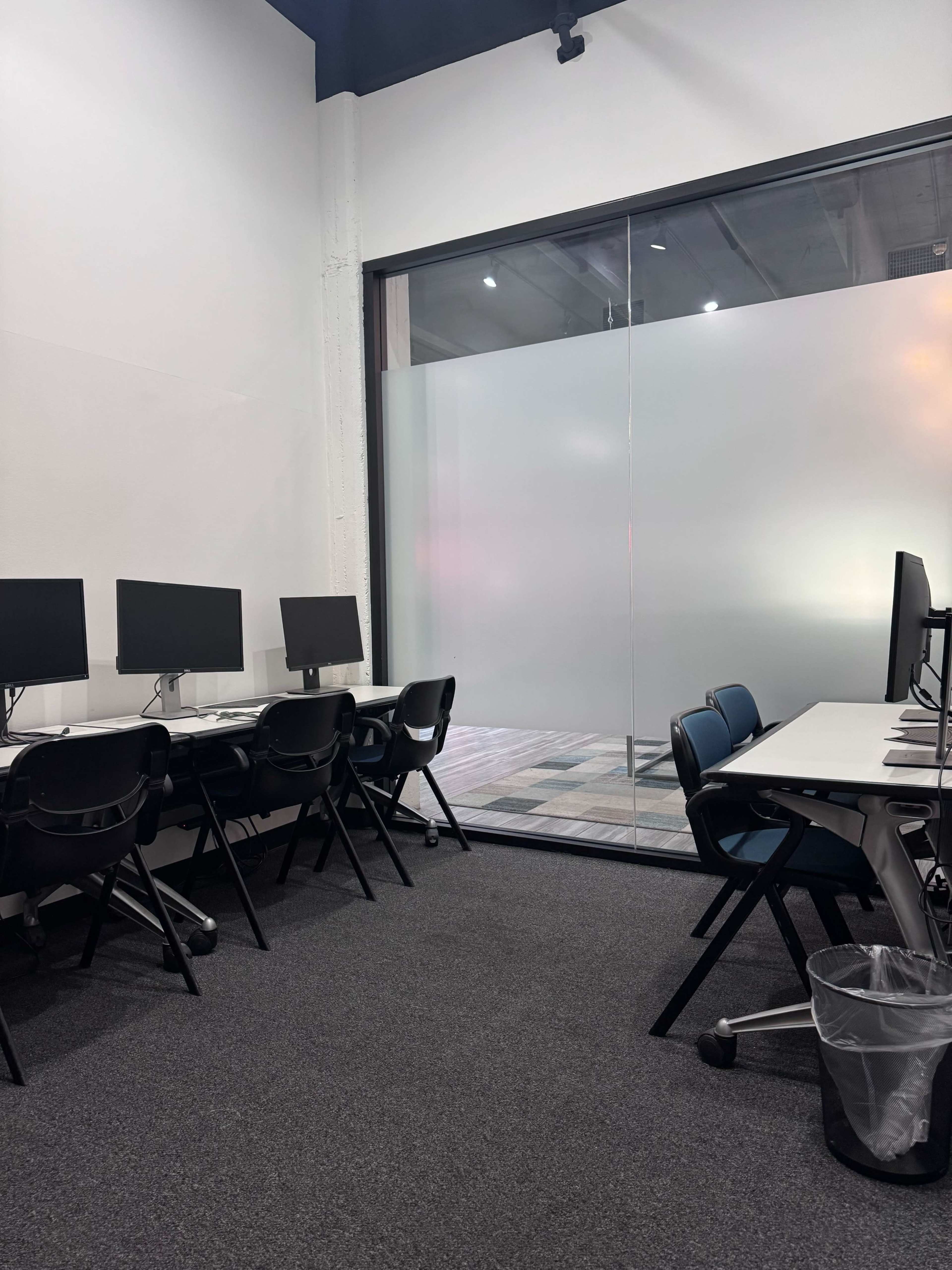 The image shows a room with several empty workstations featuring desktop computers and a glass wall partitioning a different area.