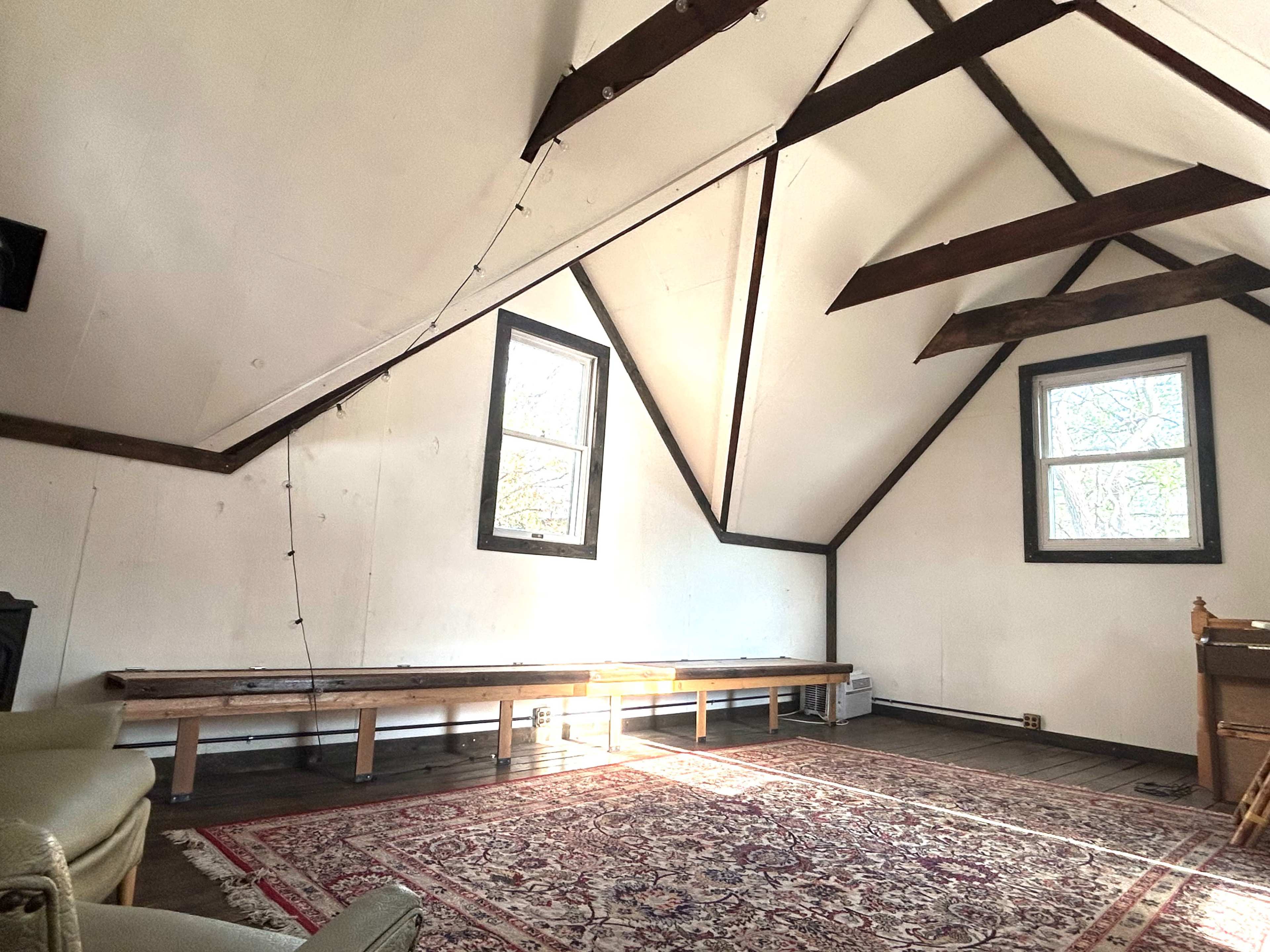 The image shows a spacious, empty room with high, slanted ceilings, two windows, and a large area rug on the wooden floor.