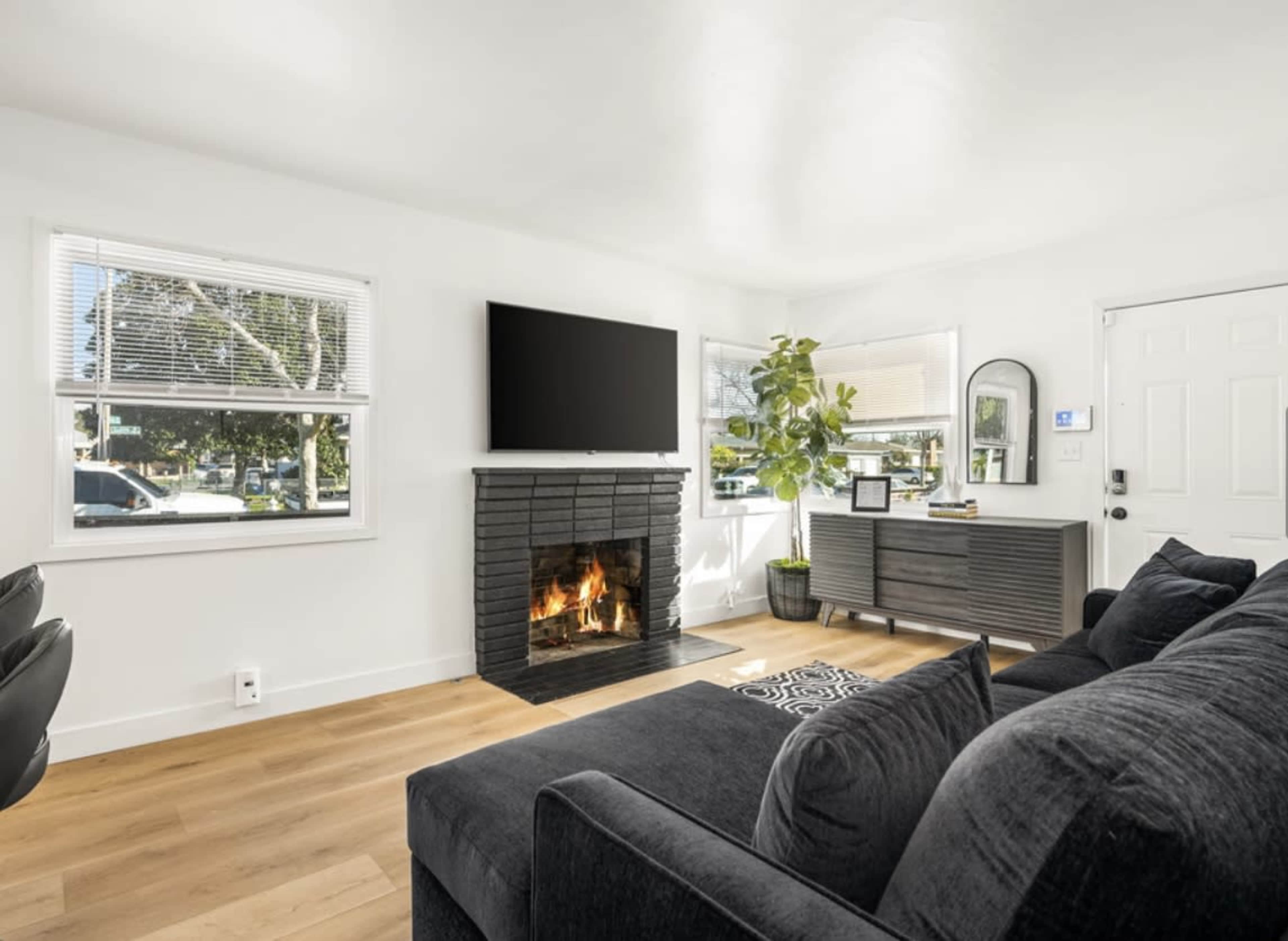 A modern living room featuring a black sofa, a fireplace with a fire burning, a television mounted on the wall, and a large window with a potted plant nearby.