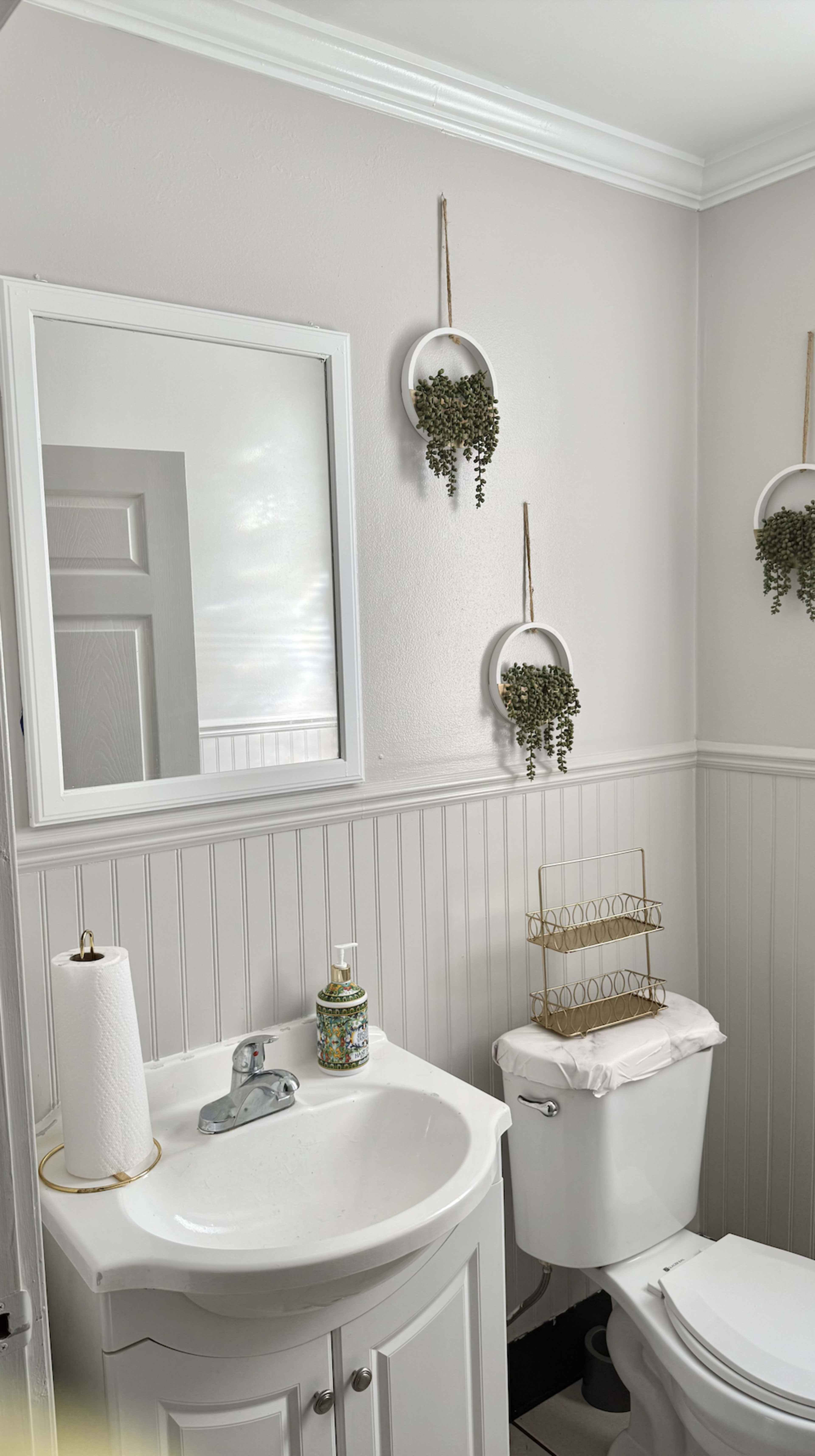 The image shows a tidy bathroom featuring a white sink with a mirror above it, a toilet beside the sink, and decorative wall planters with greenery.