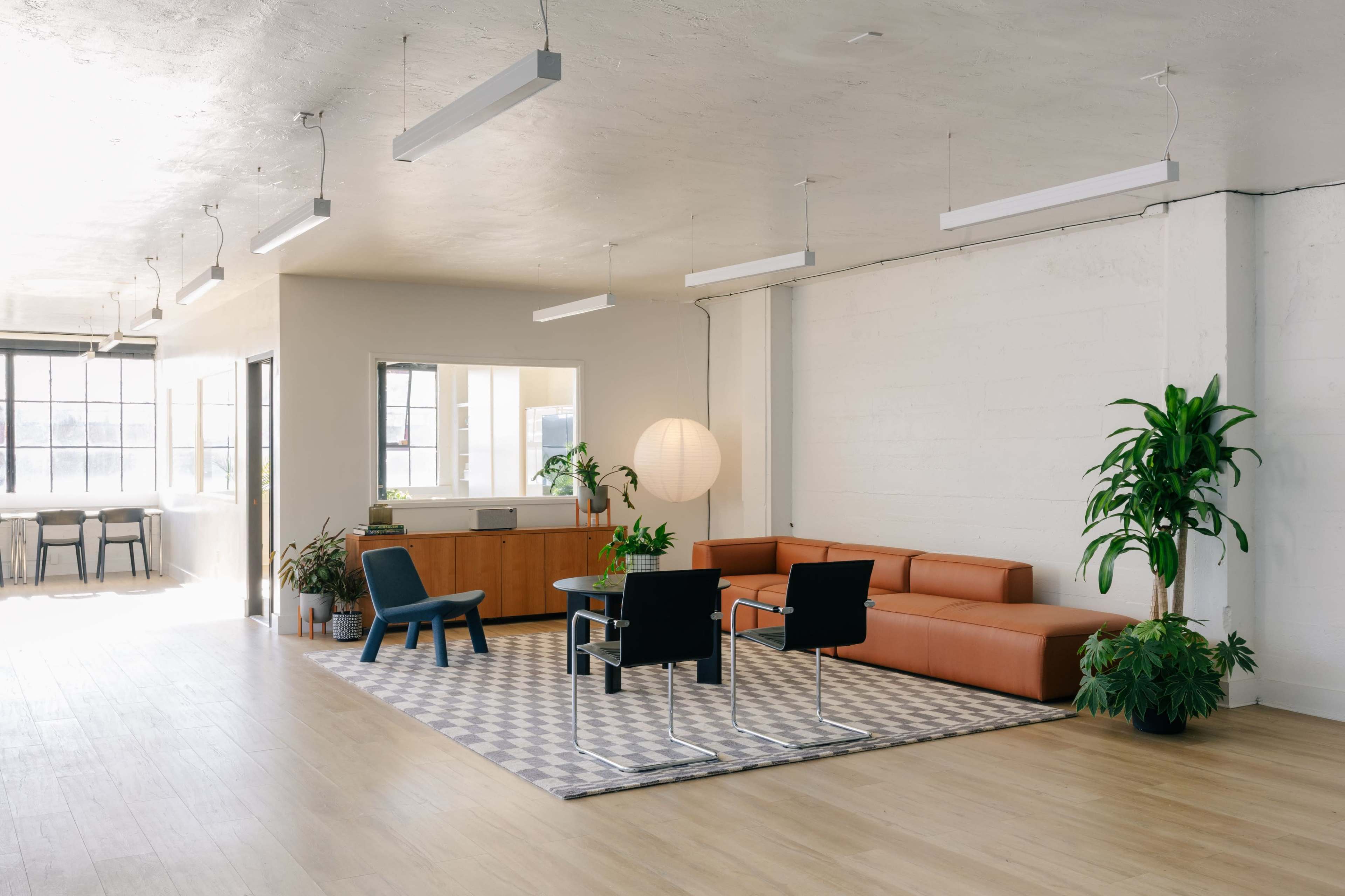The image shows a modern, spacious office interior with a brown sectional sofa, a round table surrounded by chairs, and large windows that let in natural light.