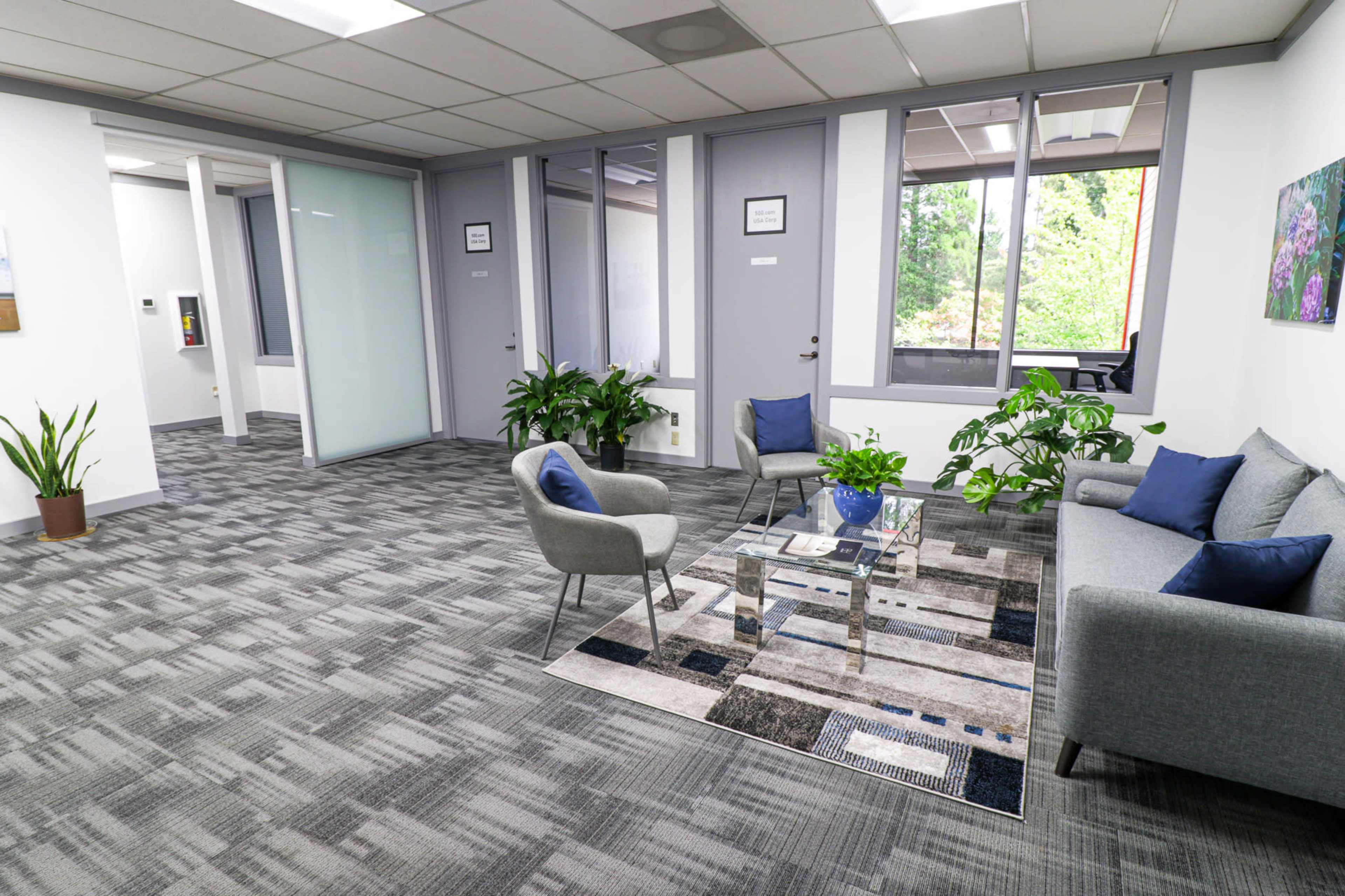 A modern office space with a seating area featuring a gray sofa, two chairs, a coffee table, and several plants, along with large windows that provide natural light.