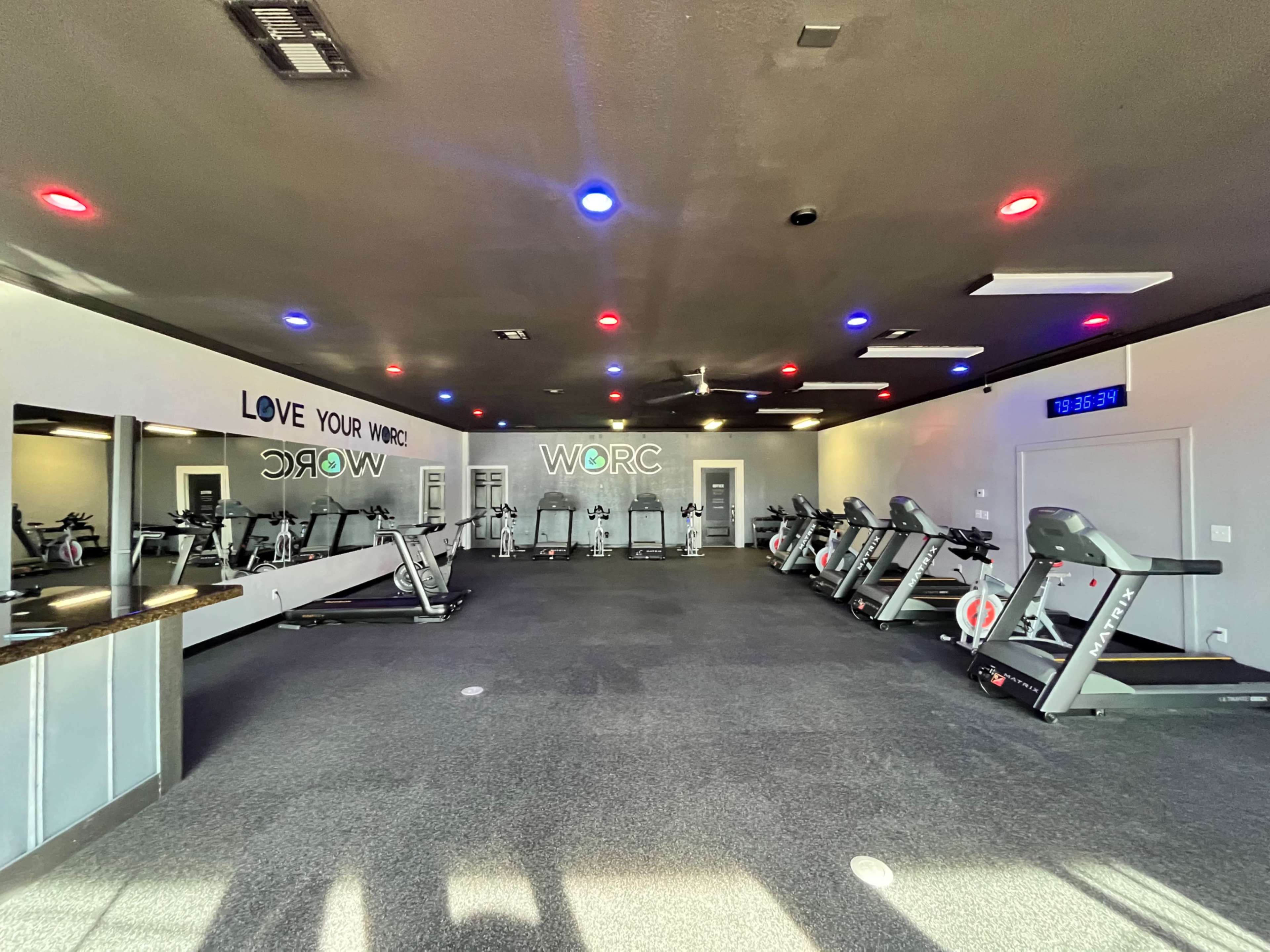 The image depicts a spacious gym interior featuring several exercise machines, mirrors on one wall, and colorful ceiling lights.
