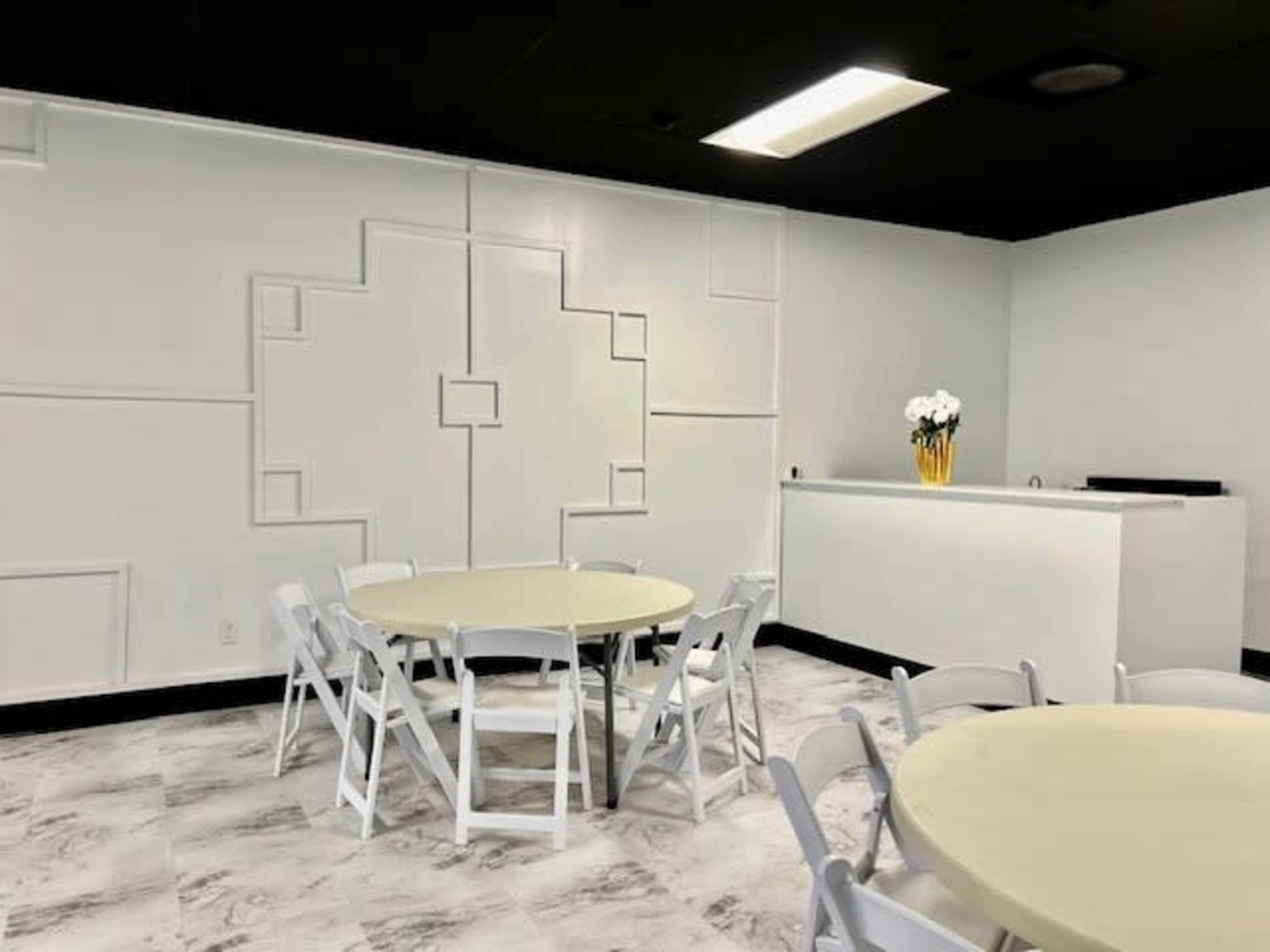 A modern room with white walls featuring geometric designs, round tables with white chairs, and a small reception area with potted flowers.