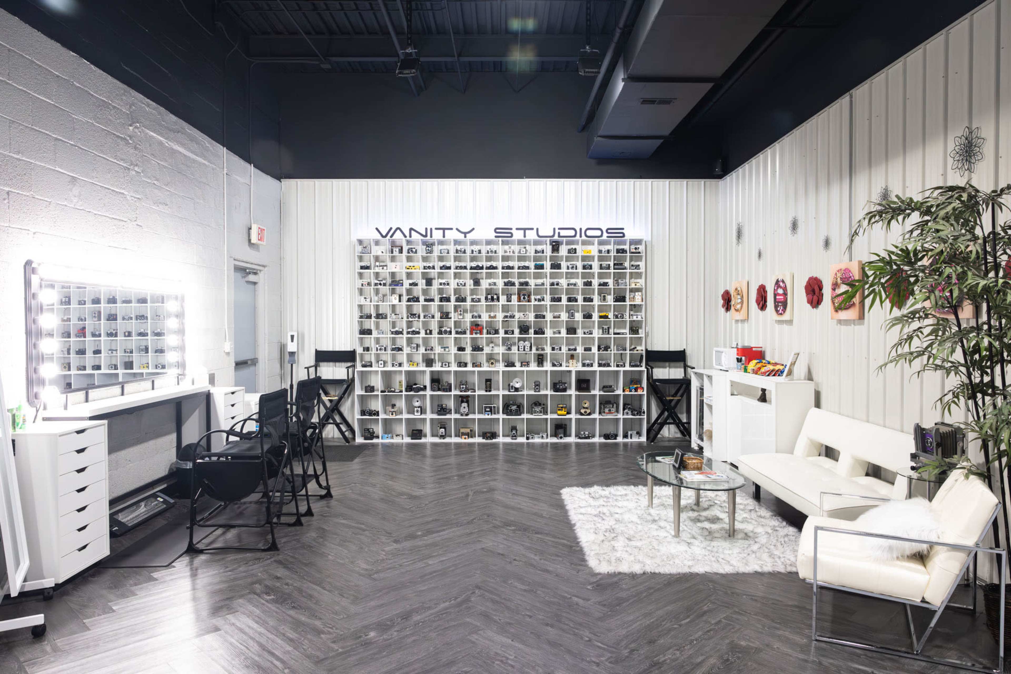 A spacious studio interior with a makeup vanity area, shelves filled with various products, and a seating area featuring a white sofa and a glass coffee table.