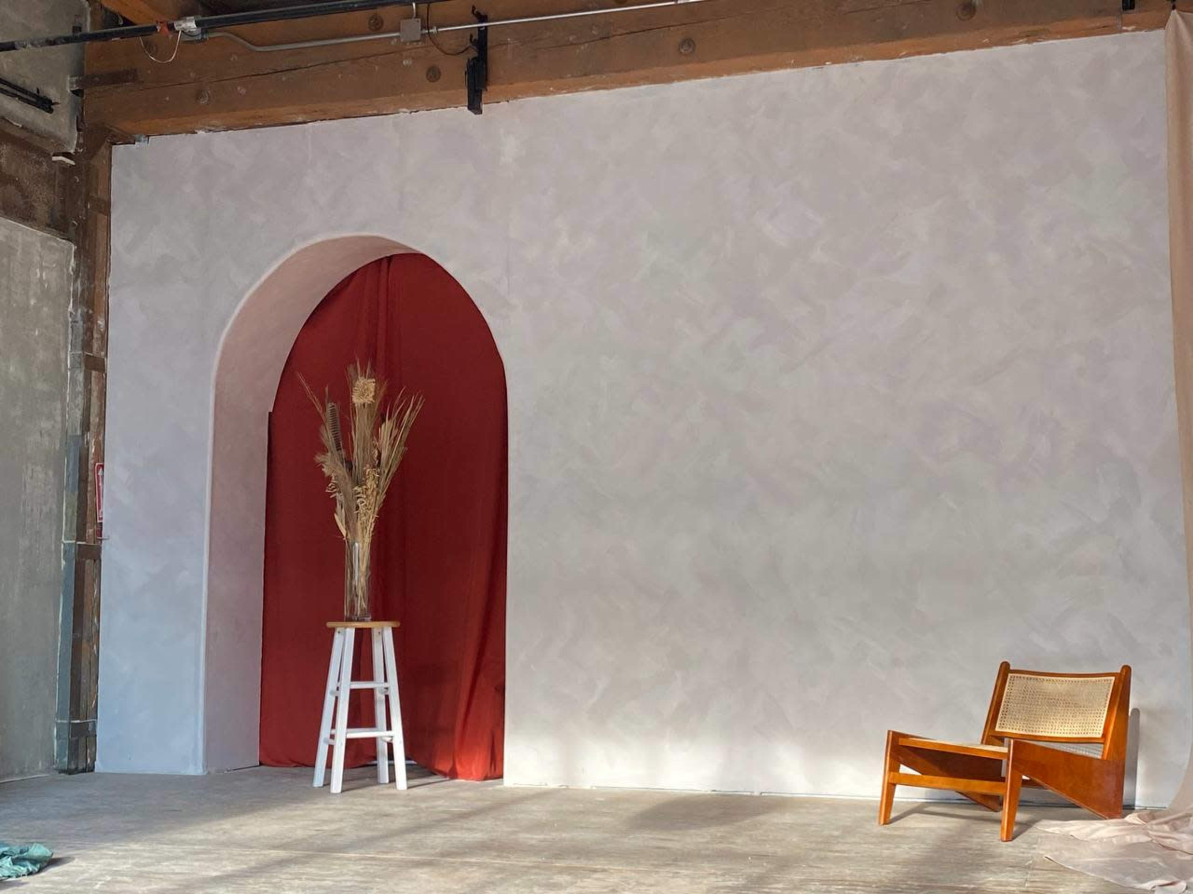 The image shows a minimalist interior space featuring a white wall with an archway covered in red fabric, a tall vase with dried plants, and a wooden chair beside a white stool.