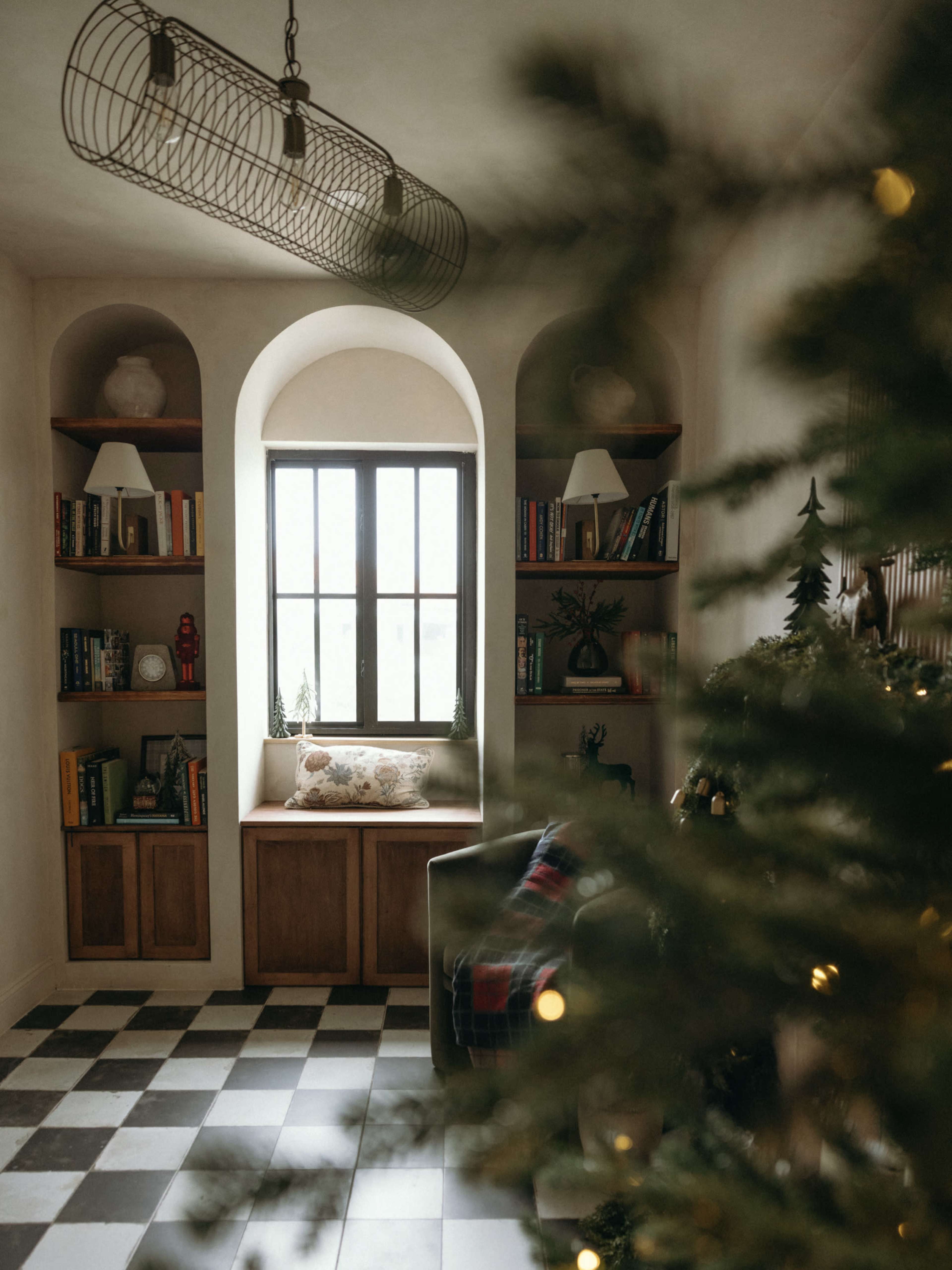 The image shows a cozy corner of a room with built-in bookshelves, a window seat, and a decorated Christmas tree.