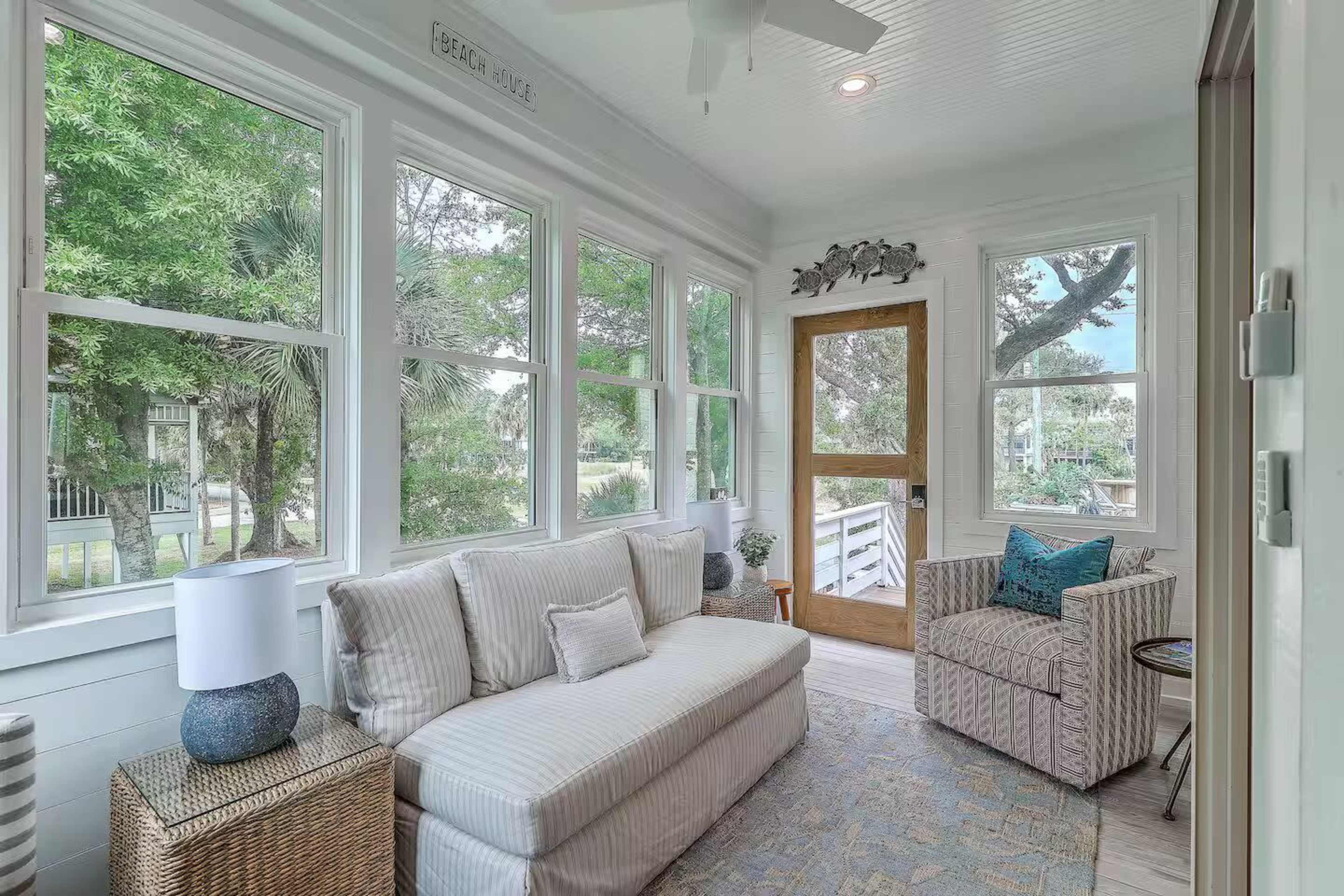 The image shows a sunlit sunroom with a white sofa, decorative pillows, a side table, and large windows overlooking greenery.