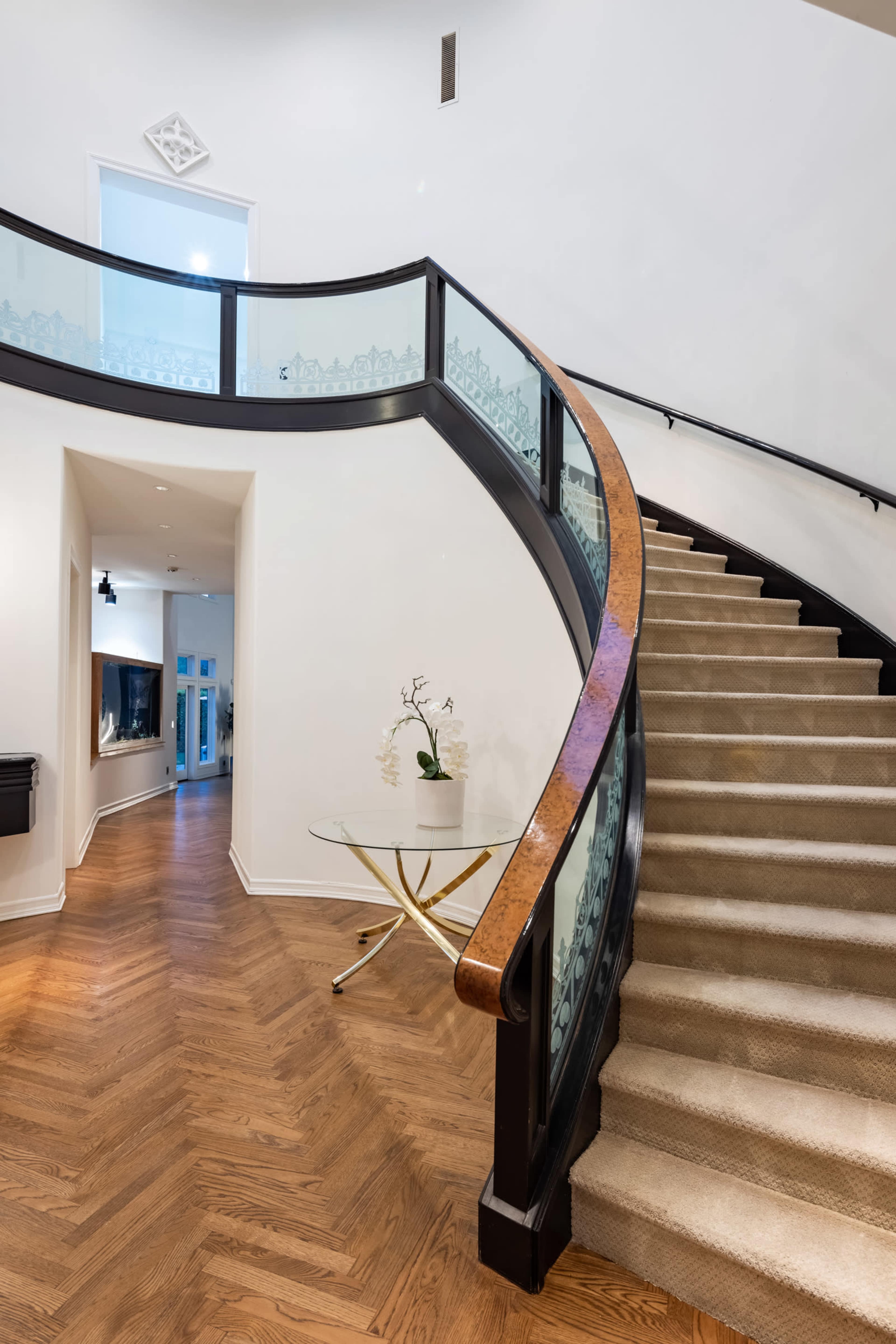 A curved staircase with a glass and wooden railing, leading to an upper level in a spacious, brightly lit entryway.