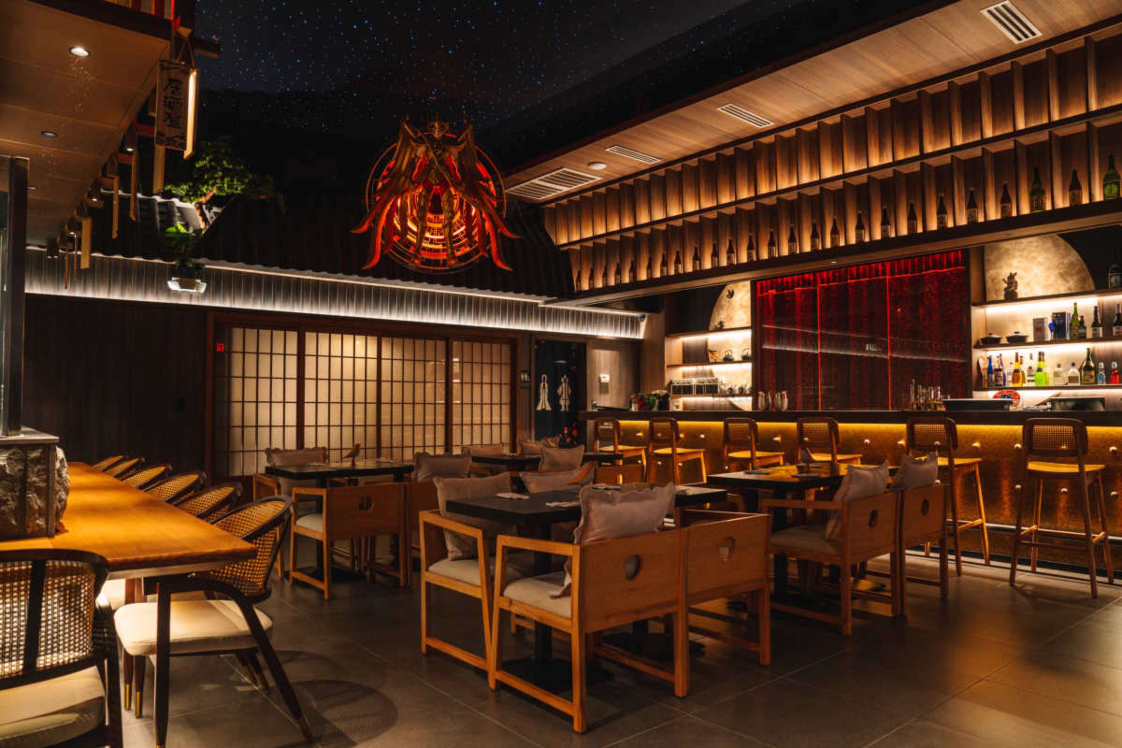 The image shows a modern restaurant interior featuring wooden furnishings, a bar with illuminated shelves, and decorative artwork on the ceiling.