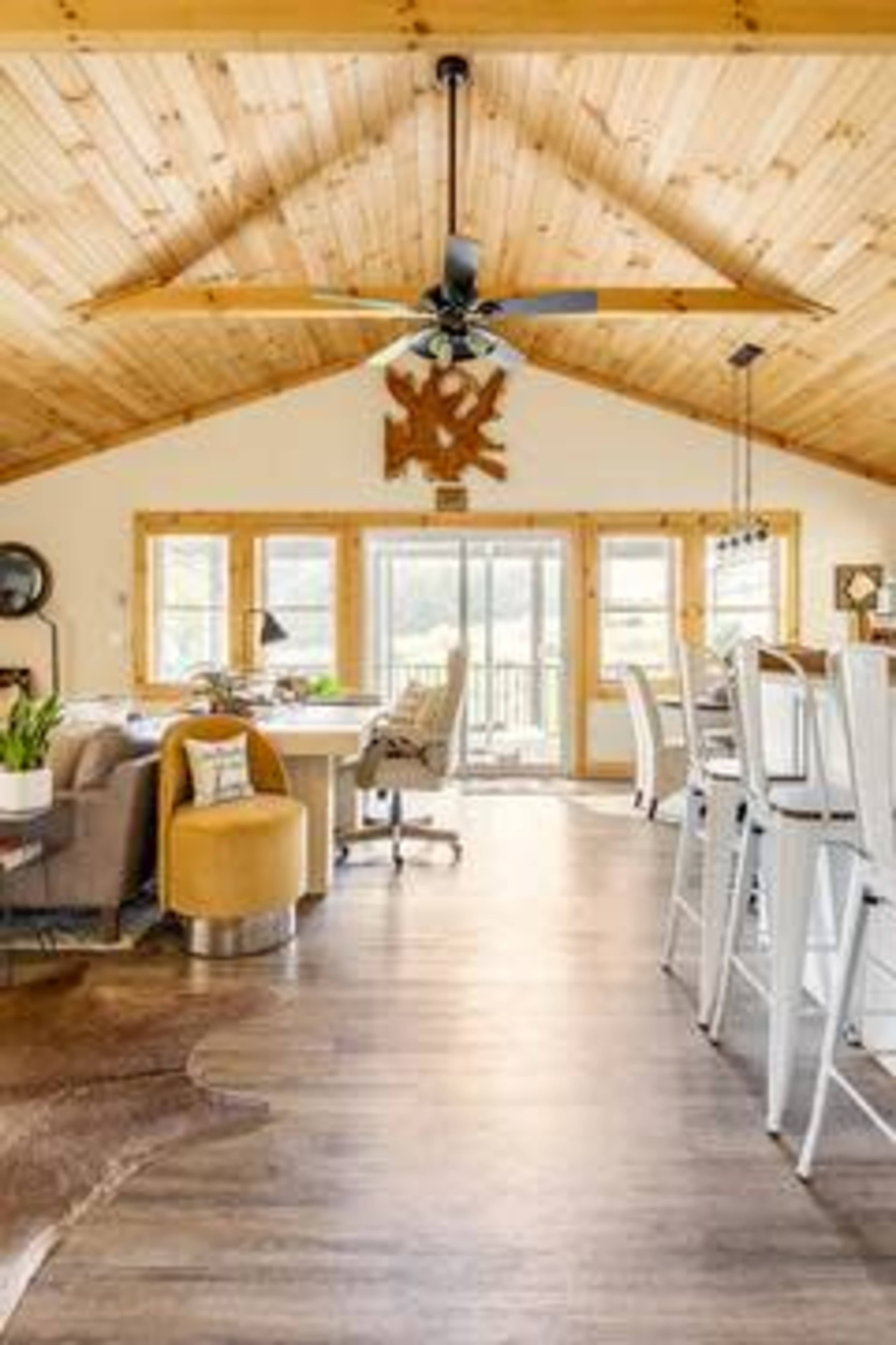 A spacious living area with a wooden ceiling, large windows, a dining table, and comfortable seating arranged around a central space.