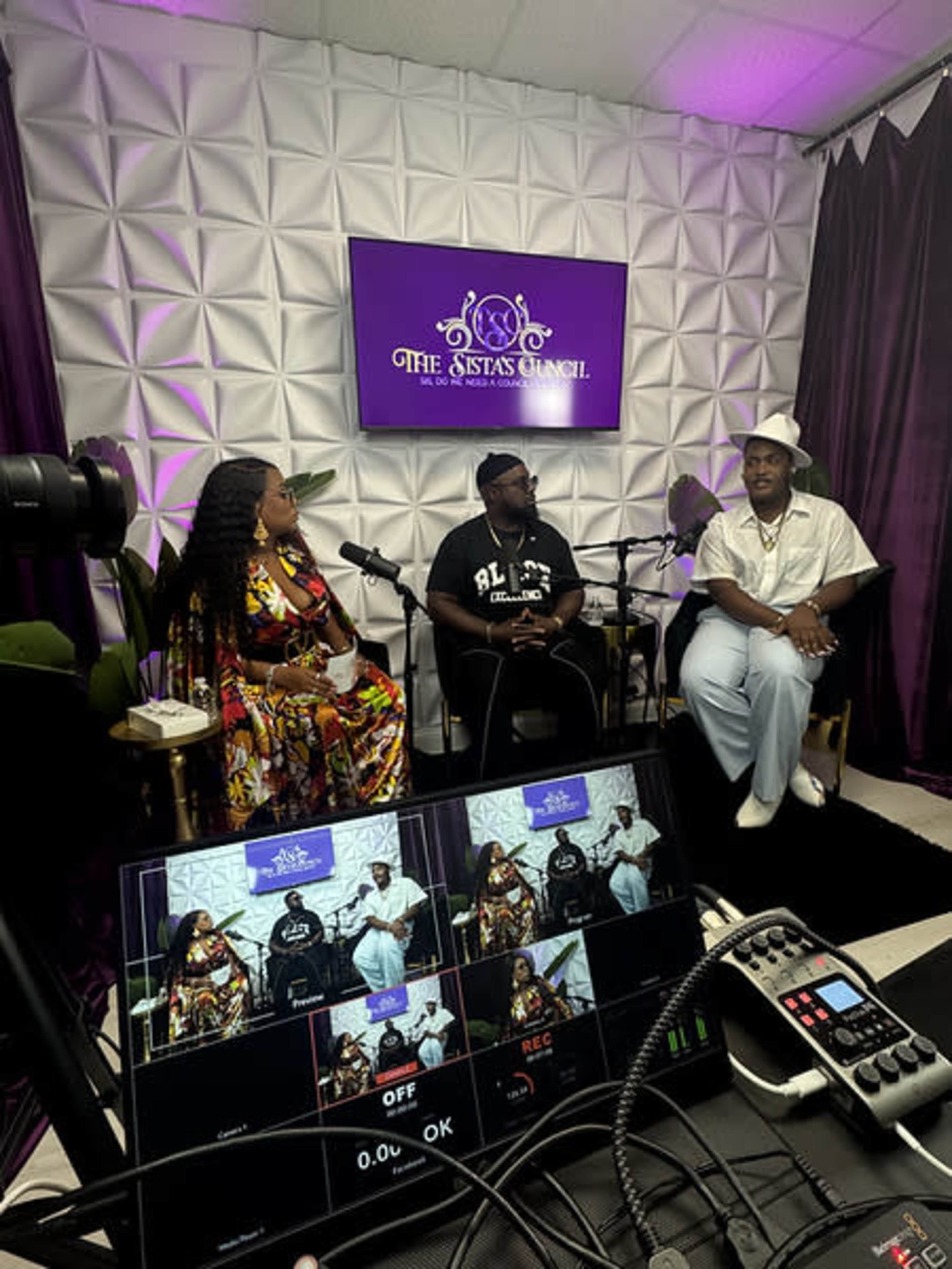A podcast recording setup features three individuals seated on a stage surrounded by purple and white decor, with microphones in front of them and equipment visible in the foreground.