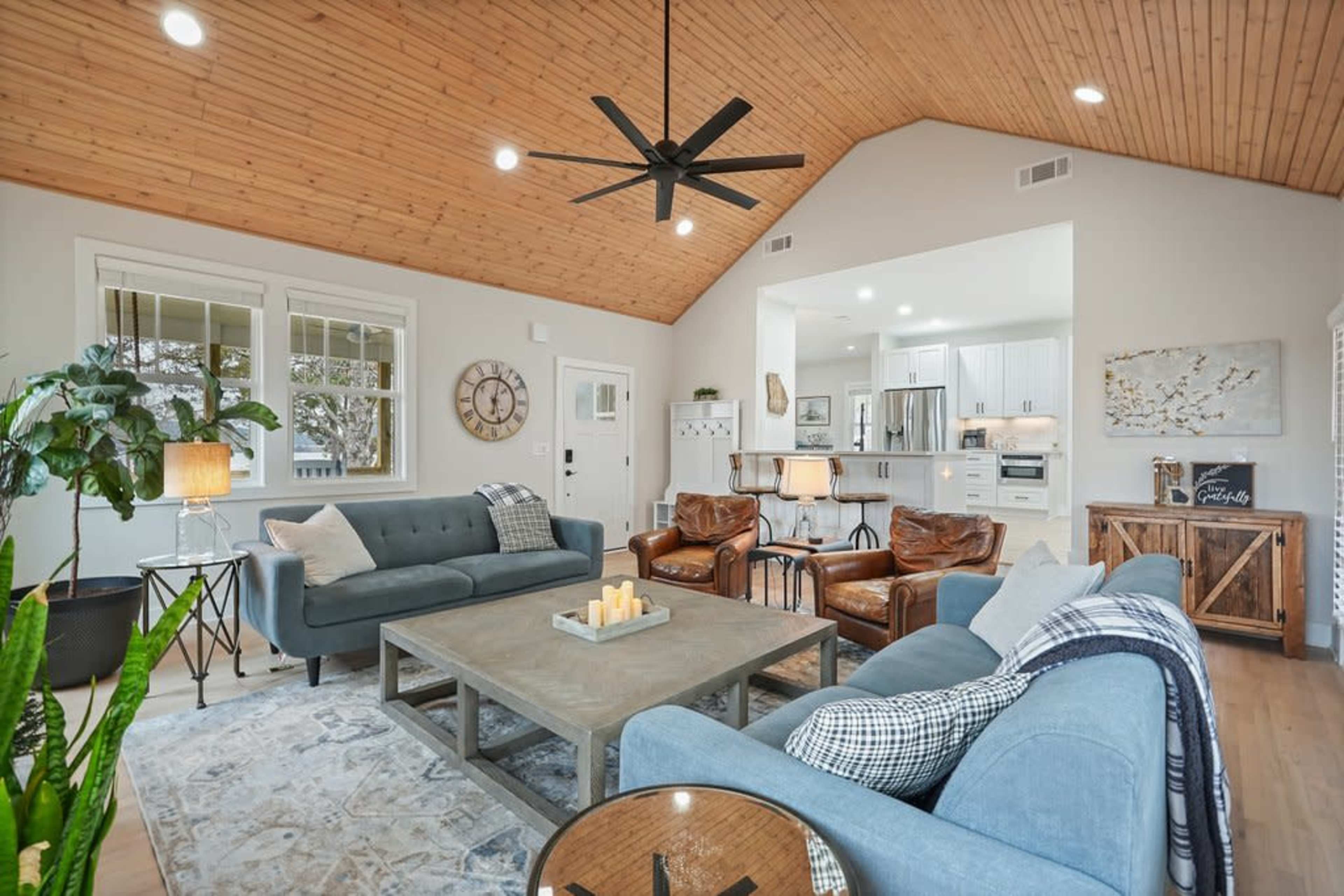 The image shows a modern living room featuring a high wooden ceiling, two sofas, leather armchairs, a coffee table with candles, and a view into an adjacent kitchen.