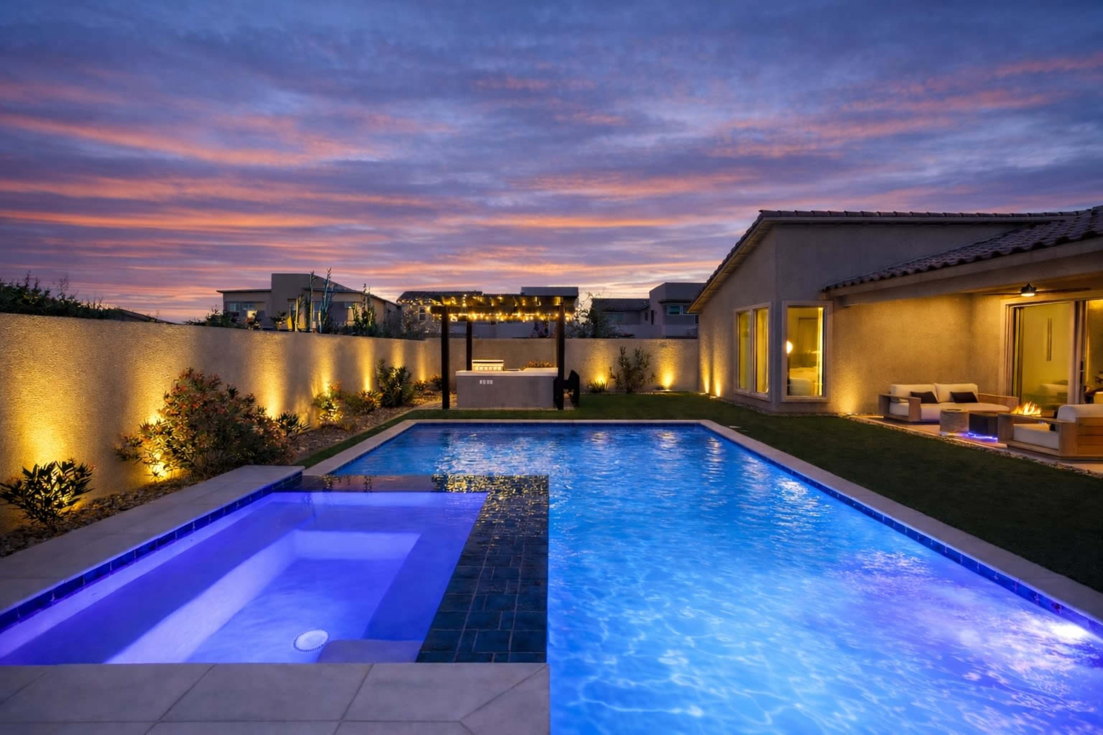 A well-lit backyard features a swimming pool and a jacuzzi, surrounded by landscaping, with a sunset sky in the background.