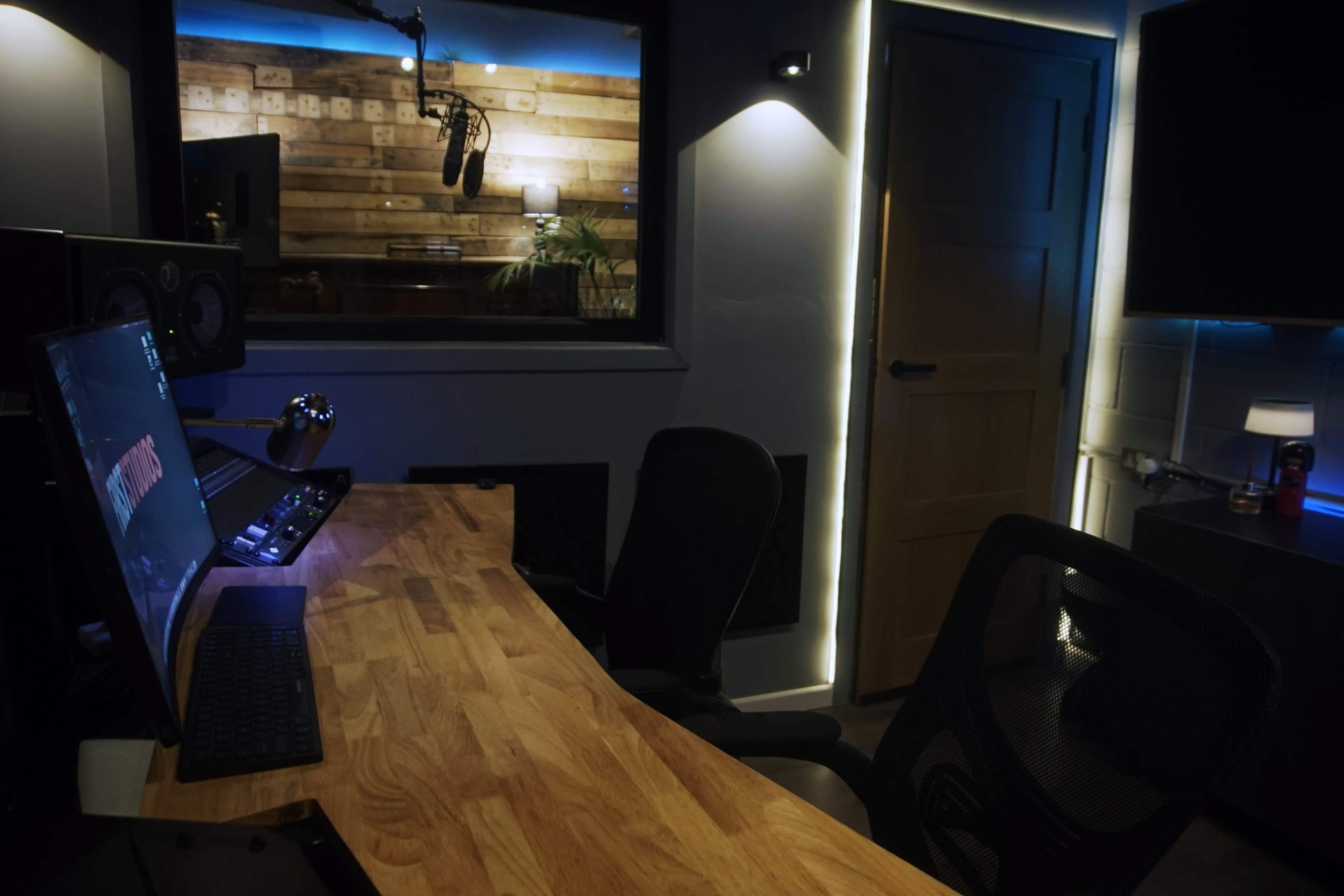 The image depicts a modern recording studio with a wooden desk, computer setup, and a microphone behind glass.