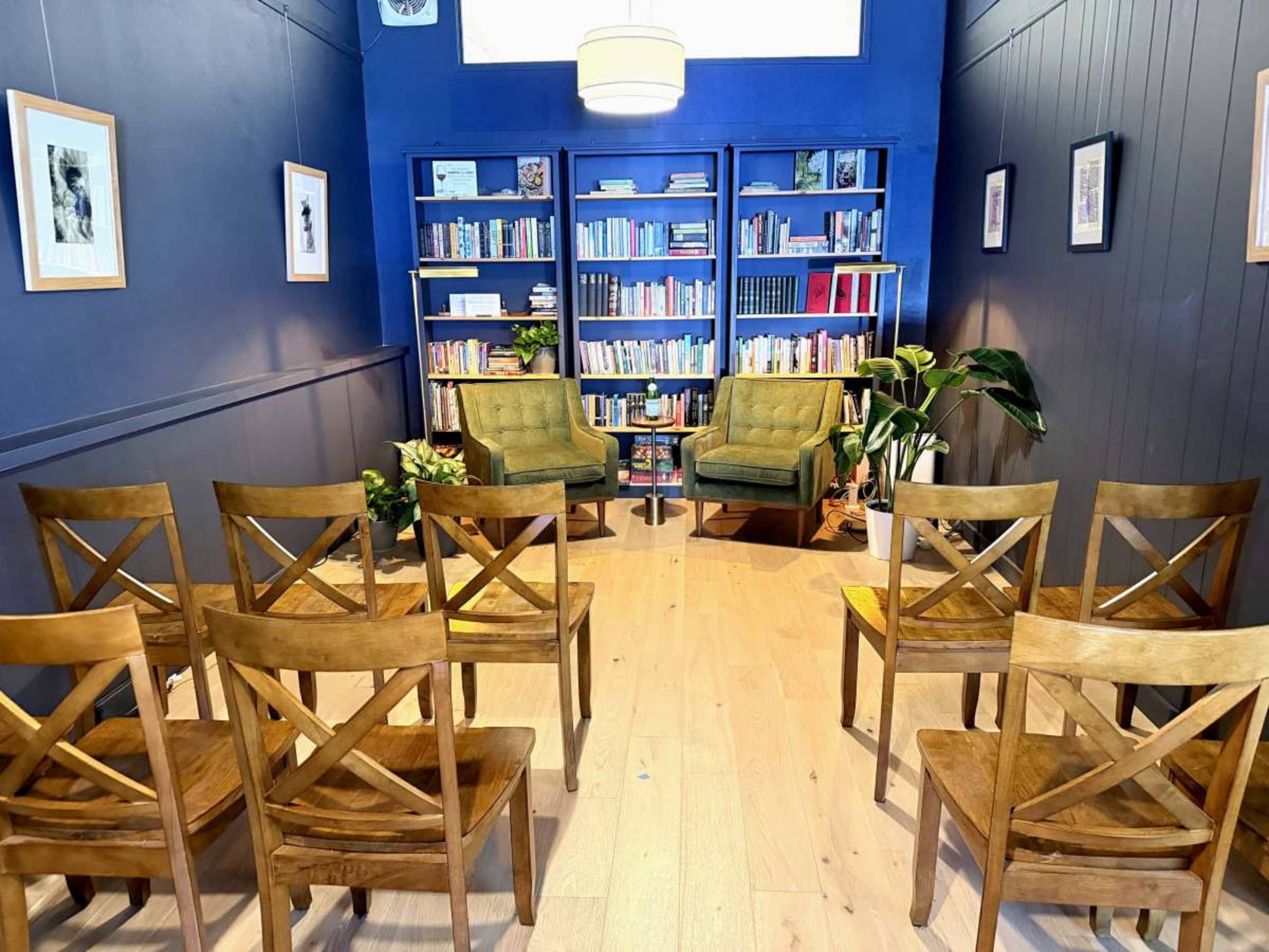 The image shows a cozy reading room with two green chairs, a bookshelf filled with books, and several wooden chairs arranged in front.