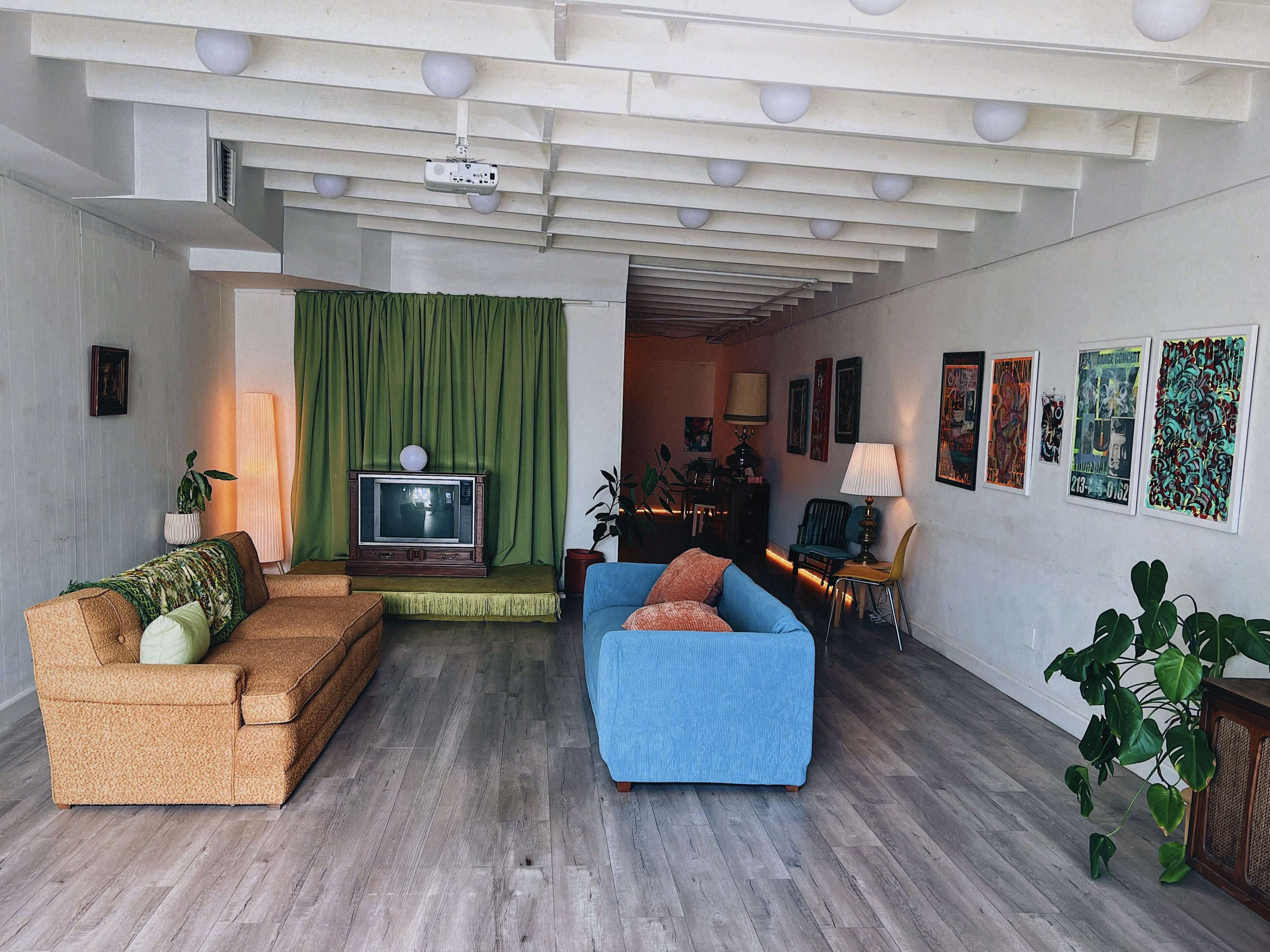 The image shows a spacious living room with two sofas, a vintage television, green curtains, and various pieces of artwork on the walls.